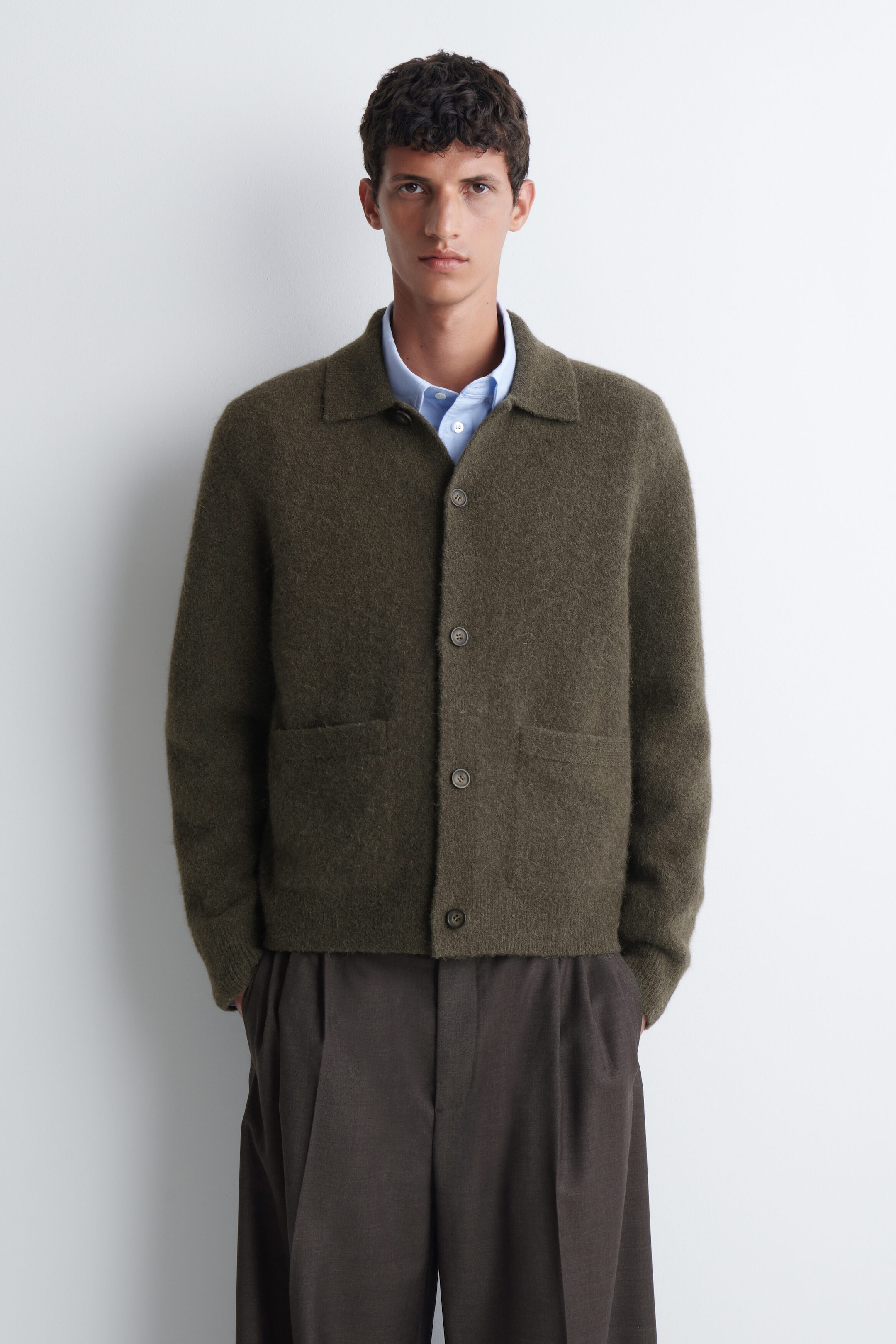 A textured collared cardigan in olive green is being worn by the model over a light blue collared shirt and dark brown trousers. The cardigan features a button-front closure, a flat collar, and front patch pockets. A textured collared cardigan in olive green is being worn by the model over a light blue collared shirt and dark brown trousers. The cardigan features a button-front closure, a flat collar, and front patch pockets.