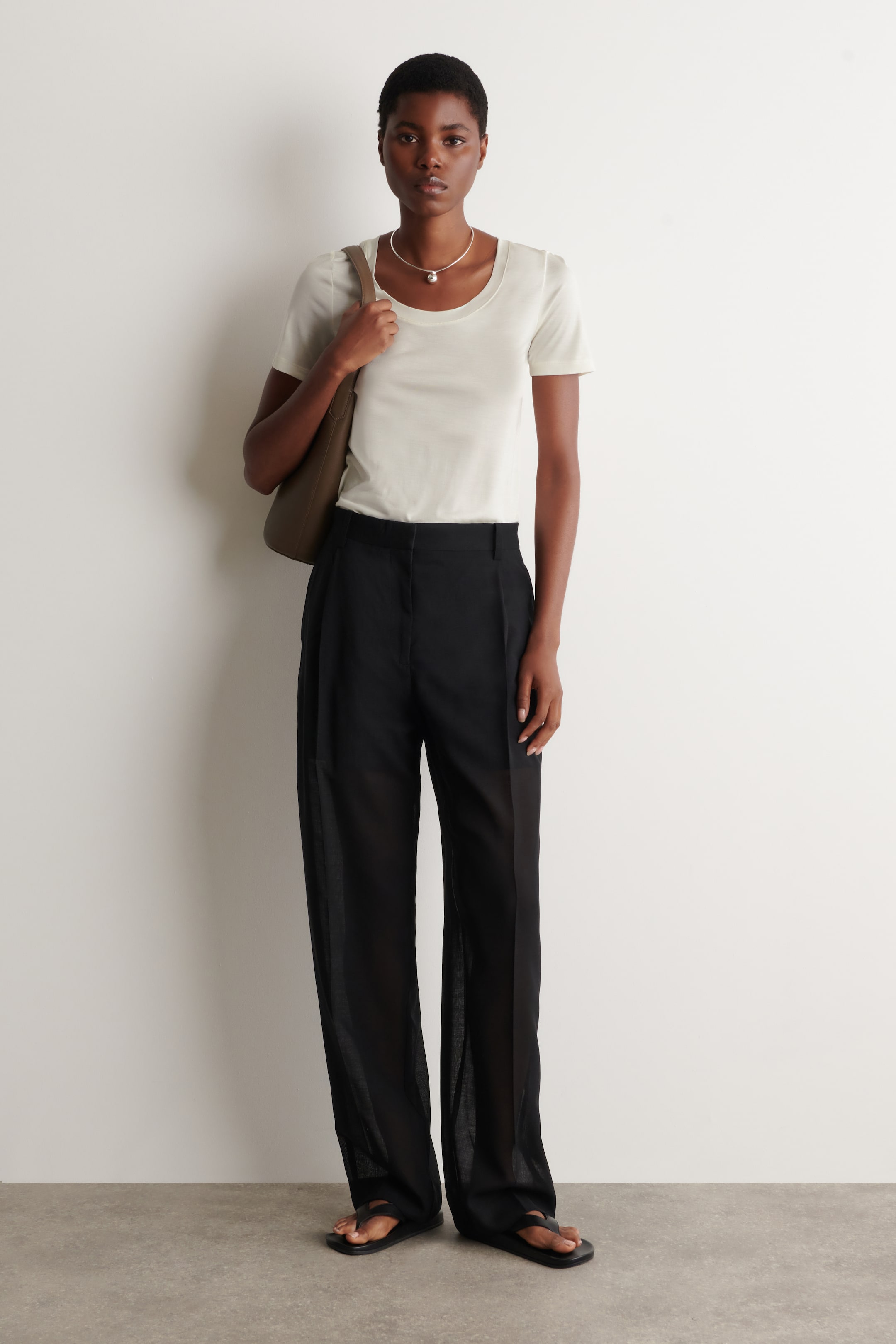 Größeres Bild ansehen: Ein elfenbeinfarbenes, kurzärmeliges T-Shirt mit einem sanft geschwungenen Ausschnitt und einer figurbetonten Silhouette wird von dem Model getragen. Es ist in eine schwarze Hose gesteckt und mit einer silbernen Halskette sowie einer braunen Umhängetasche kombiniert.