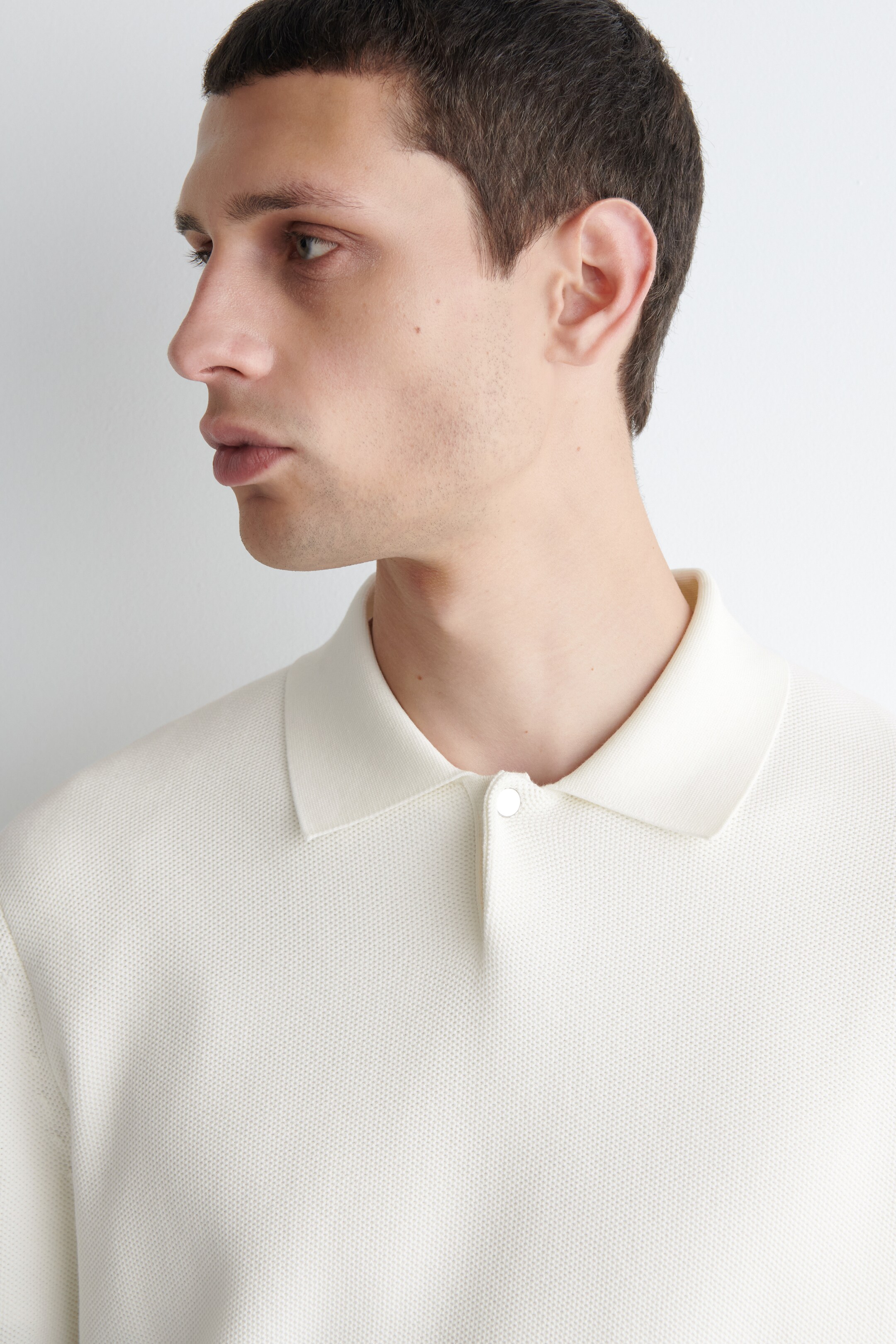 View larger image: A cream-colored polo shirt is being worn, its textured pique knit fabric is showing. Its classic collar and a partial placket with one white button are being displayed from a side profile.