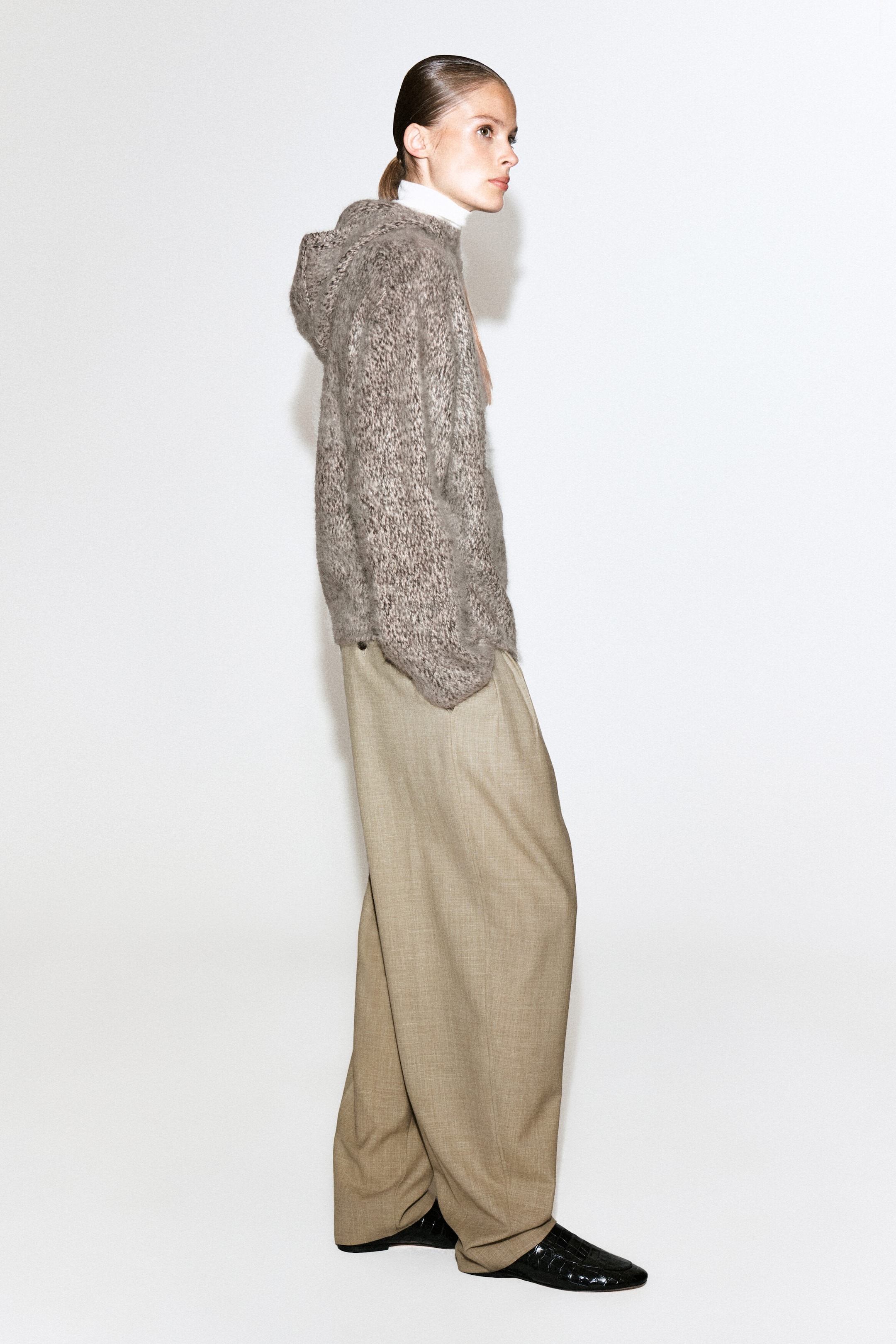 A chunky brushed-cashmere hoodie in a textured, heathered brown knit is worn by the model over a white high-neck top. The hoodie is styled with wide-leg beige trousers and black shoes, with the model shown in side profile. A chunky brushed-cashmere hoodie in a textured, heathered brown knit is worn by the model over a white high-neck top. The hoodie is styled with wide-leg beige trousers and black shoes, with the model shown in side profile.
