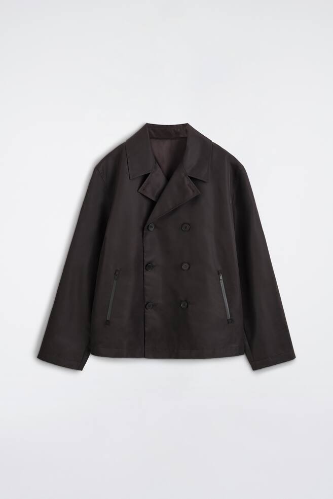 Een technische korte trenchcoatjas van nylon in donkerbruin wordt van voren getoond, met een dubbele rij knopen met zes knopen en klassieke revers met inkeping. De jas heeft een korte, boxy silhouet met twee verticale handzakken met ritssluiting en netjes afgewerkte manchetten. - 1