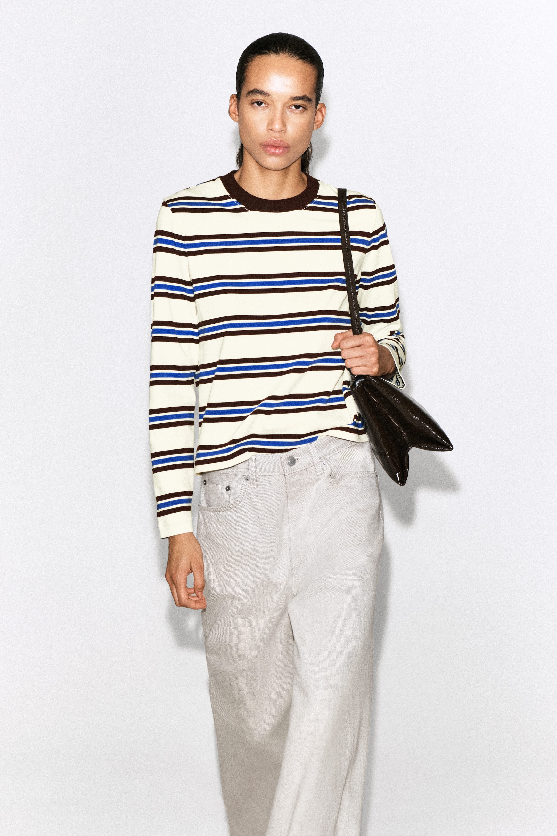 A clean-cut long-sleeved T-shirt is shown from the front, featuring horizontal stripes in cream, dark brown and bright blue. The garment has a solid dark brown ribbed crew neck and straight-cut long sleeves, and falls loosely over the waistband of light-coloured trousers. A clean-cut long-sleeved T-shirt is shown from the front, featuring horizontal stripes in cream, dark brown and bright blue. The garment has a solid dark brown ribbed crew neck and straight-cut long sleeves, and falls loosely over the waistband of light-coloured trousers.