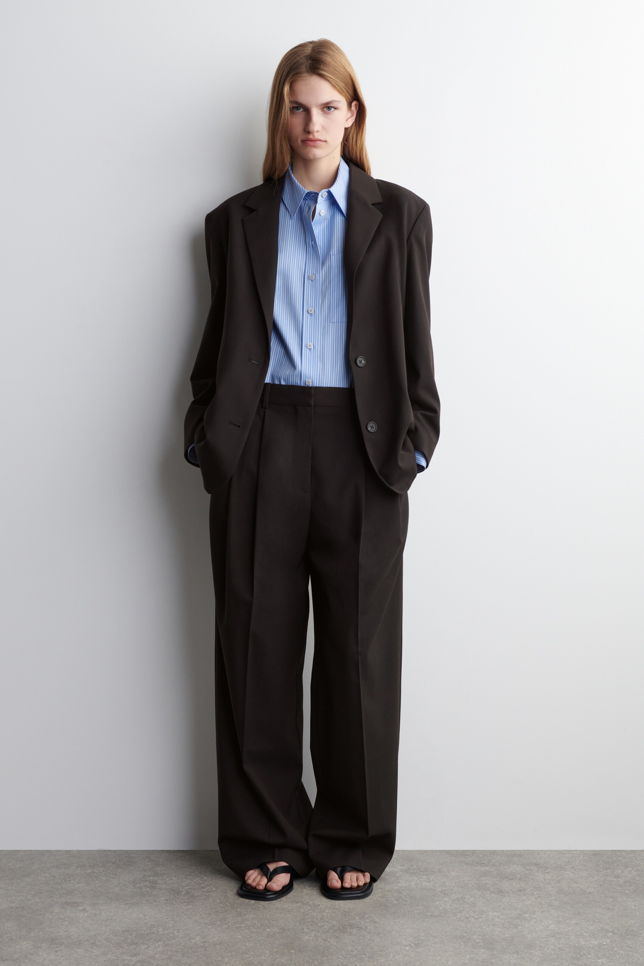 Größeres Bild anzeigen: Ein dunkelbrauner, übergroßer Anzug wird in einer Frontalansicht präsentiert, mit einem Blazer mit breiten Schultern, Kerbrevers und zwei vorderen Knöpfen, zusammen mit passenden, weit geschnittenen Hosen mit Frontfalten. Ein hellblau-weiß gestreiftes Button-Down-Hemd wird unter dem Blazer getragen, und schwarze Sandalen mit offenen Zehen werden getragen.