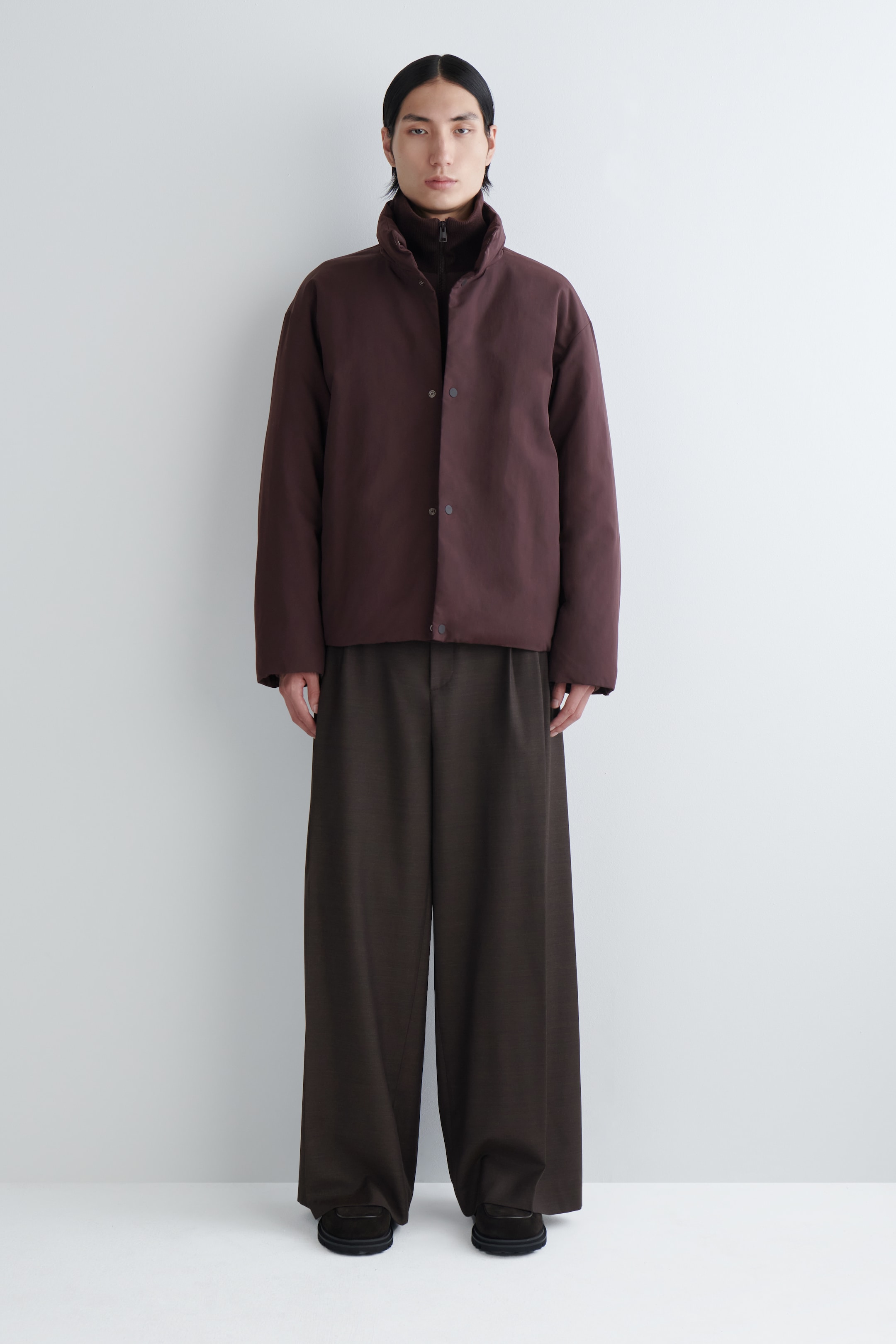 View larger image: A dark brown, relaxed-fit short jacket is being presented from a front angle, featuring a high stand collar and visible front snap buttons, layered over a dark high-neck top. Dark brown wide-leg pants are being styled with the jacket, pooling slightly at the ankles over dark slip-on shoes.