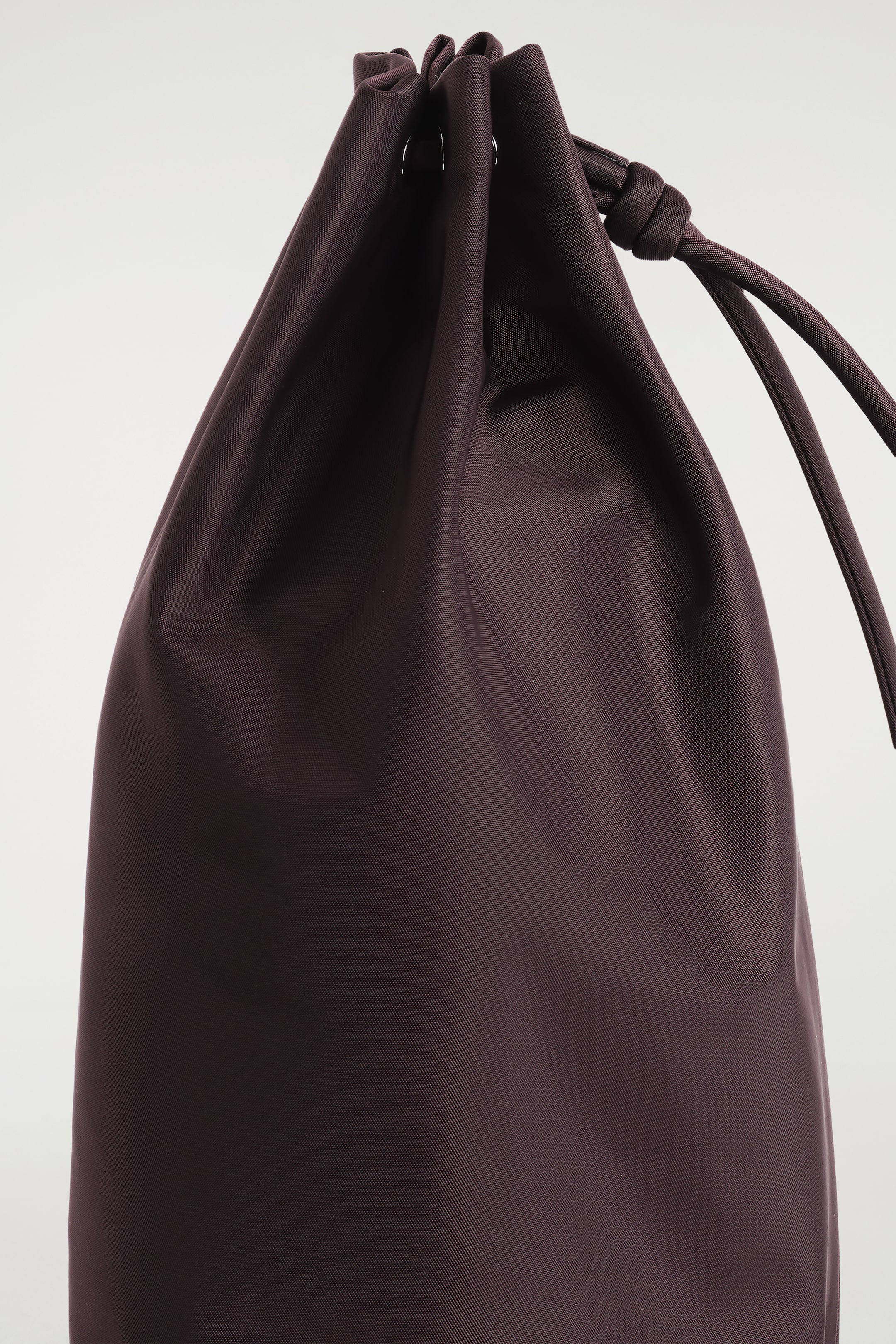 View larger image: A dark plum-brown drawstring bag is standing upright, angled slightly to the right, with its top cinched closed by a matching drawstring that is knotted on the right side. Two silver-toned eyelets are visible on the left, and the fabric is displaying a subtle diagonal texture.