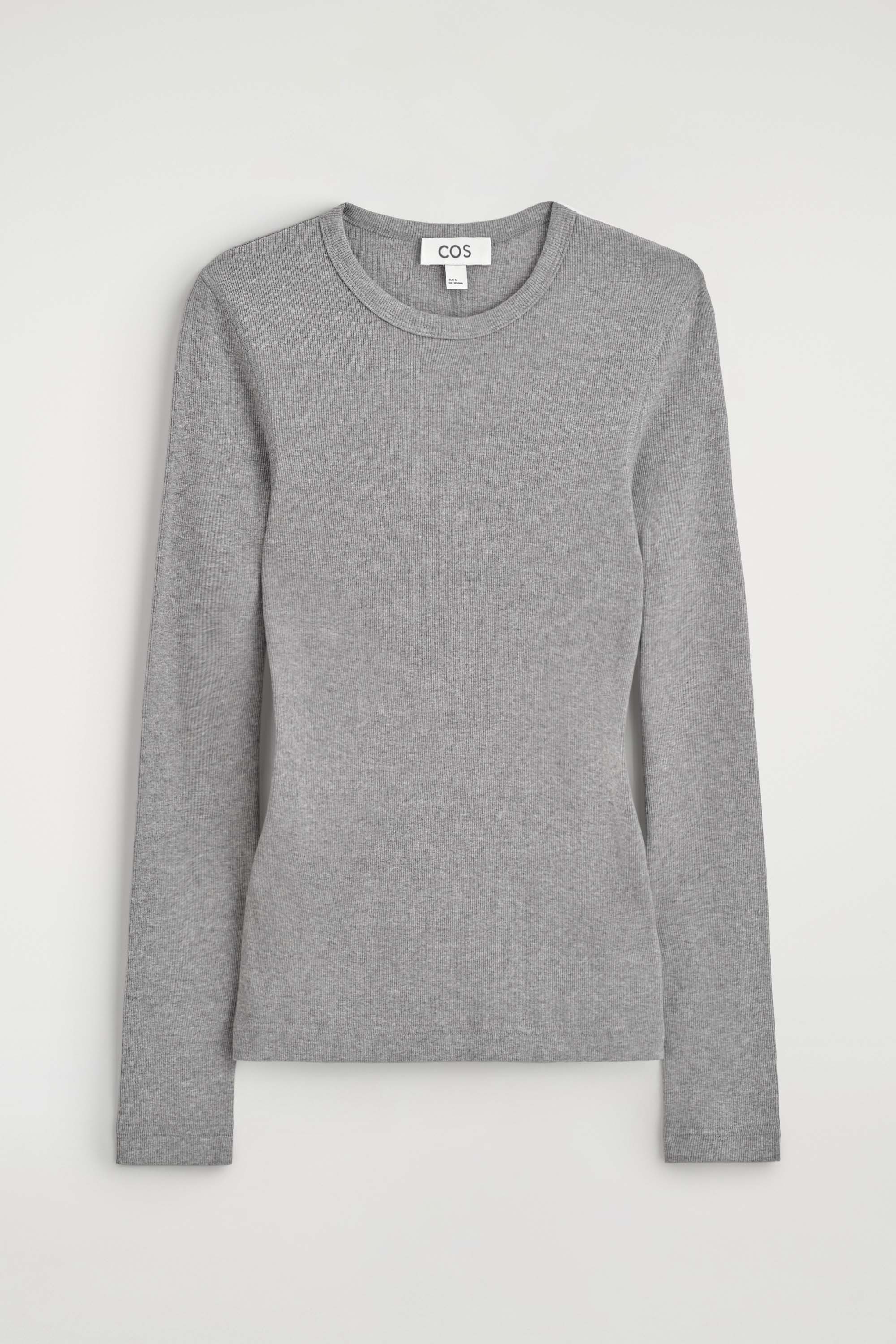 The slim ribbed cotton long-sleeved top is shown from the front in a close-fitting silhouette, featuring a light grey colour, a classic crew neck, and a fine ribbed texture. The top rests untucked at the hips over a black skirt, emphasising its slim-fit design.