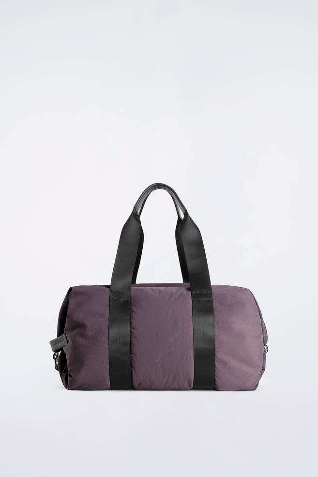 Le sac de voyage en nylon, dâun violet sourd, est prÃ©sentÃ© debout de face, mettant en valeur sa forme cylindrique et ses deux larges sangles noires en toile qui lâenserrent verticalement. Ces sangles se prolongent vers le haut pour former des poignÃ©es supÃ©rieures finies avec des dÃ©tails en cuir noir. - 1