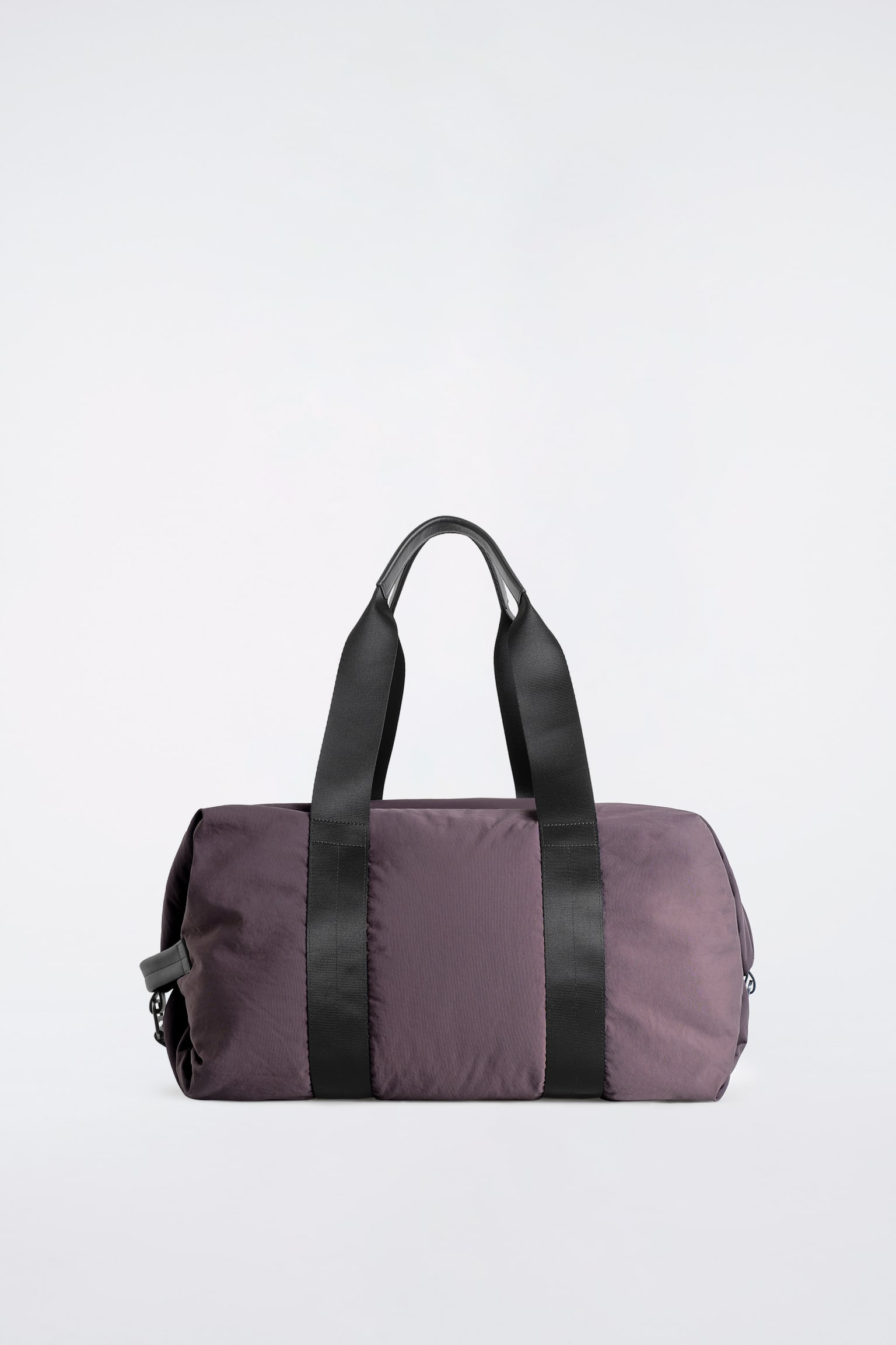 Le sac de voyage en nylon à détails en cuir est présenté de face, mettant en valeur sa structure cylindrique souple dans une teinte taupe-brun foncé. Il est doté de deux larges poignées supérieures en sangle noire avec des prises gainées de cuir qui descendent verticalement le long du corps, ainsi que dâune petite tirette en cuir sur le côté. - 1