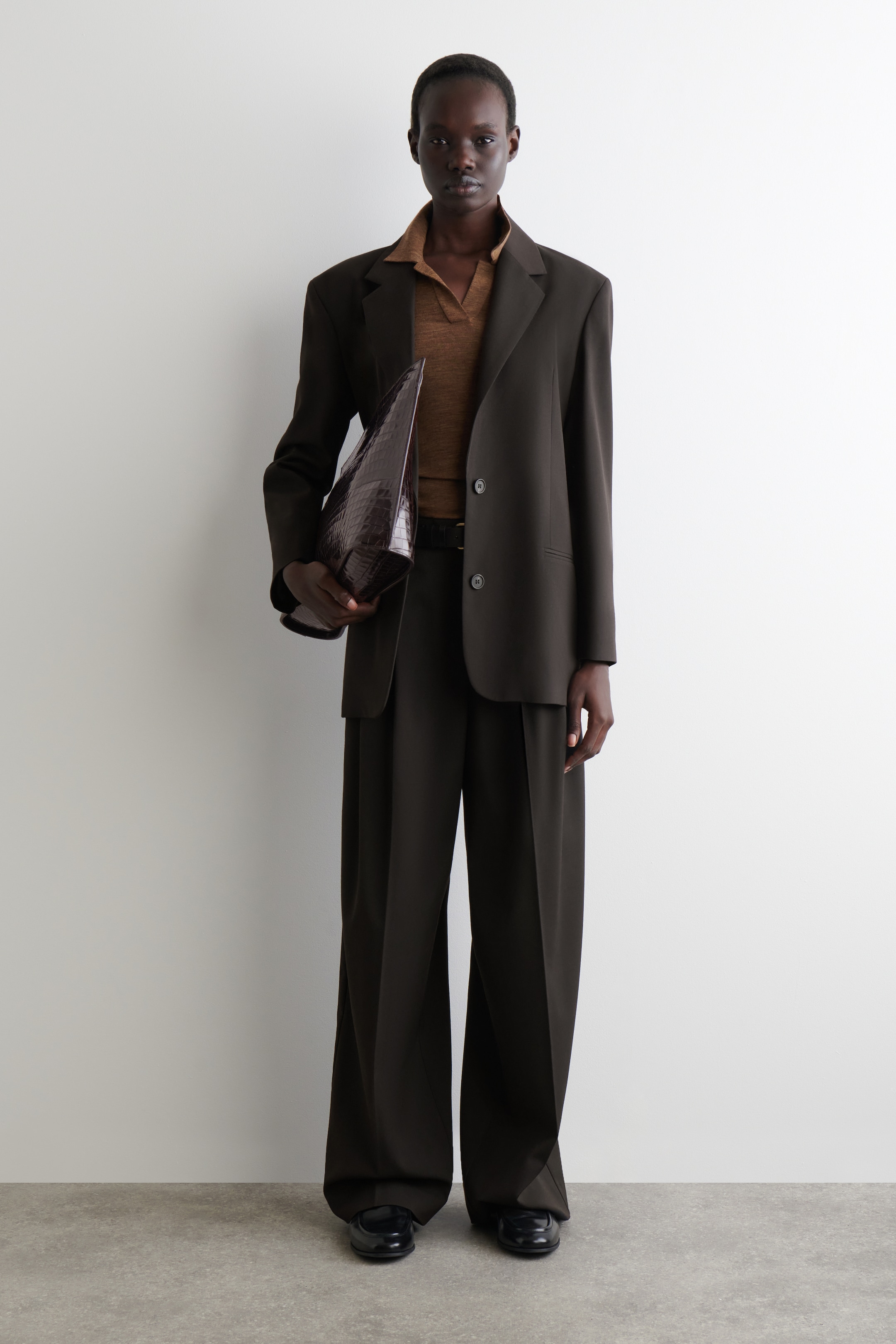 Visualizza immagine più grande: Un abito marrone scuro, caratterizzato da una giacca oversize con revers a tacca e pantaloni a gamba larga con pieghe, è indossato. Una polo marrone a maglia è stratificata sotto, e una borsa tote marrone scuro con stampa coccodrillo è tenuta in mano, mentre delle scarpe nere in stile mocassino piatto sono indossate.