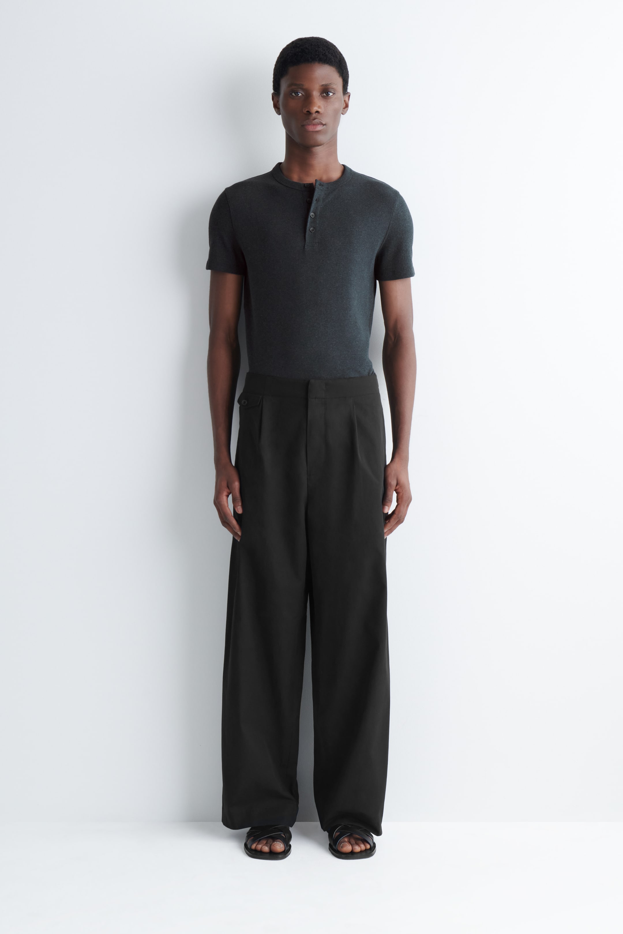 Größeres Bild ansehen: Schwarze, weit geschnittene Hosen mit großzügigen Frontfalten, Schnallen an der Taille und einer Münztasche werden vom Model getragen. Das Model kombiniert die Hose mit einem dunklen Kurzarmhemd und schwarzen Sandalen.