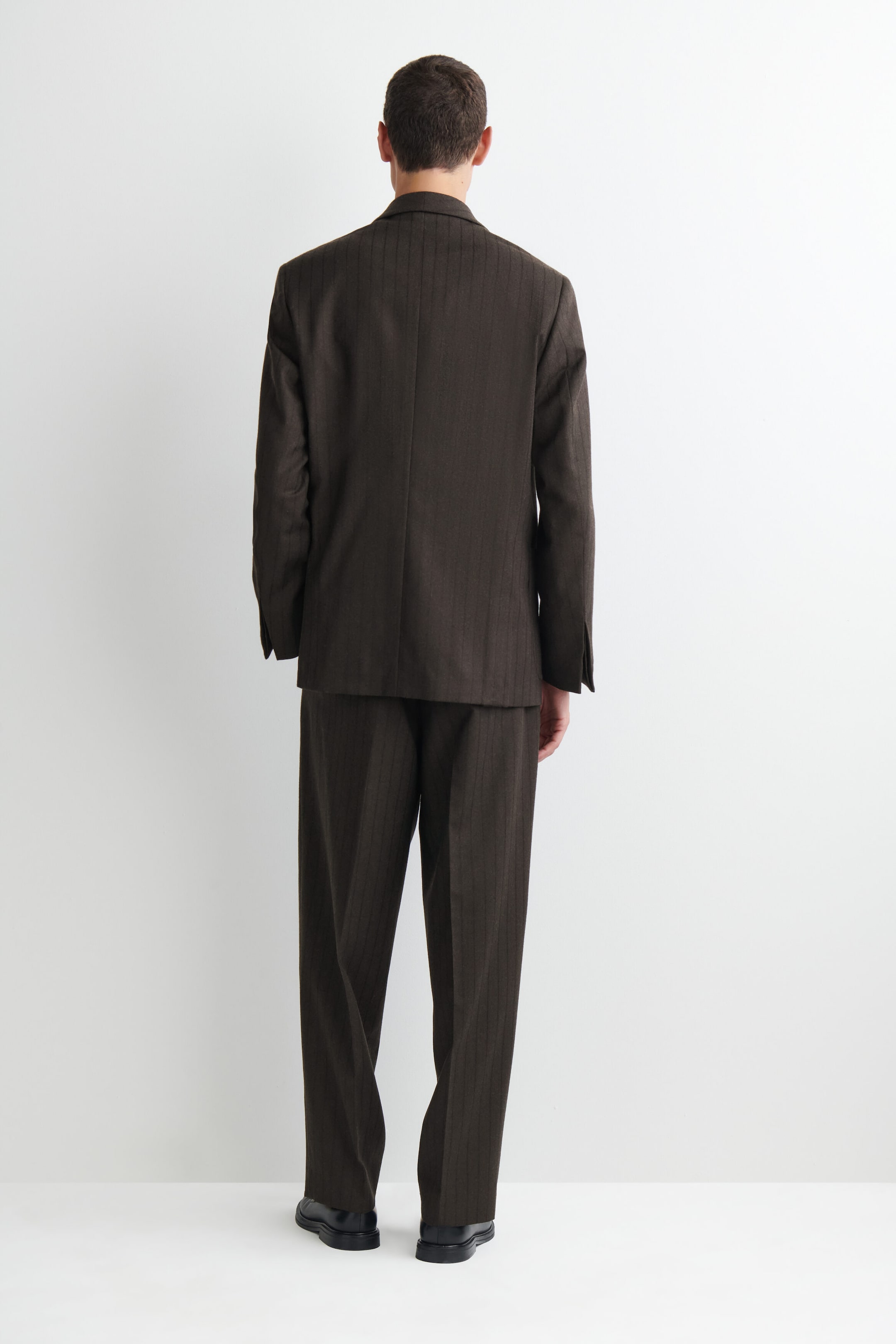 A model is wearing a relaxed-fit, dark brown pinstripe wool blazer and matching trousers, facing away to showcase the suit's clean drape and structured shoulders. A model is wearing a relaxed-fit, dark brown pinstripe wool blazer and matching trousers, facing away to showcase the suit's clean drape and structured shoulders.