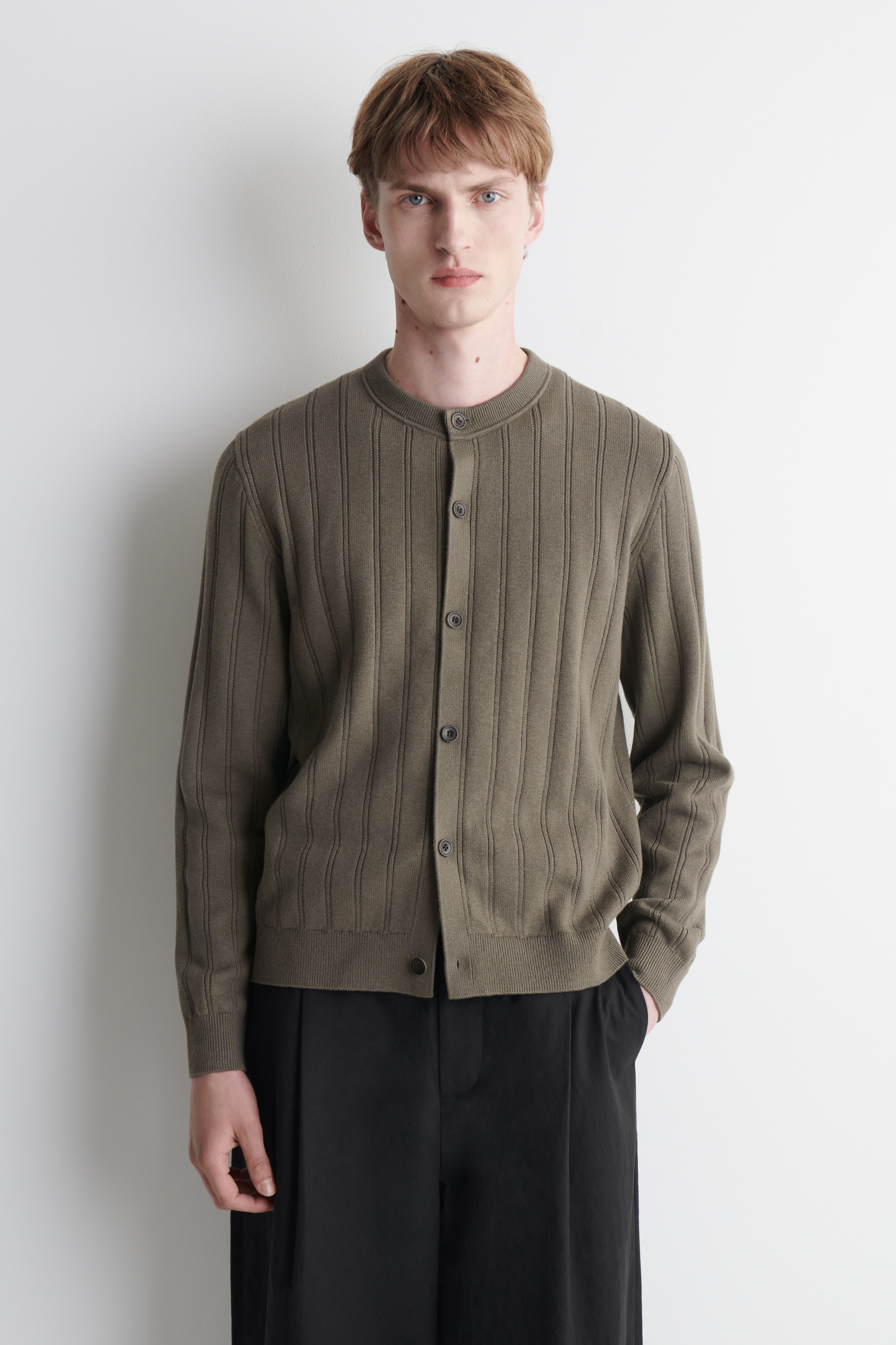An olive green cardigan is shown from the front, featuring a ribbed knit texture throughout the body and sleeves. It has a crew neck, five dark buttons down the centre front, ribbed cuffs, and a ribbed hem.