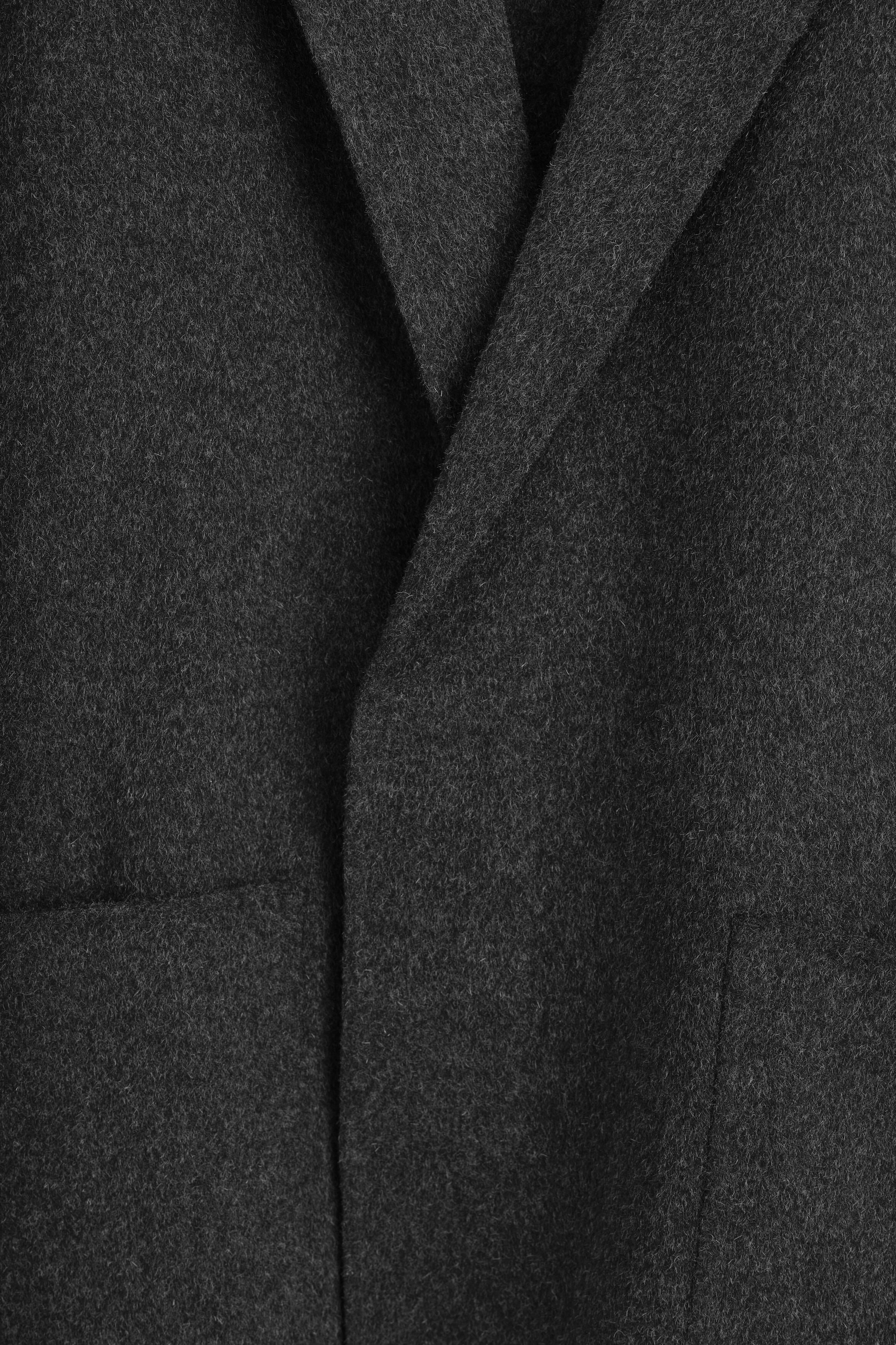 A close-up of a dark grey wool-blend blazer showing the soft, textured surface of the double-faced fabric and the clean, unstructured finish of the lapel and front opening. A close-up of a dark grey wool-blend blazer showing the soft, textured surface of the double-faced fabric and the clean, unstructured finish of the lapel and front opening.