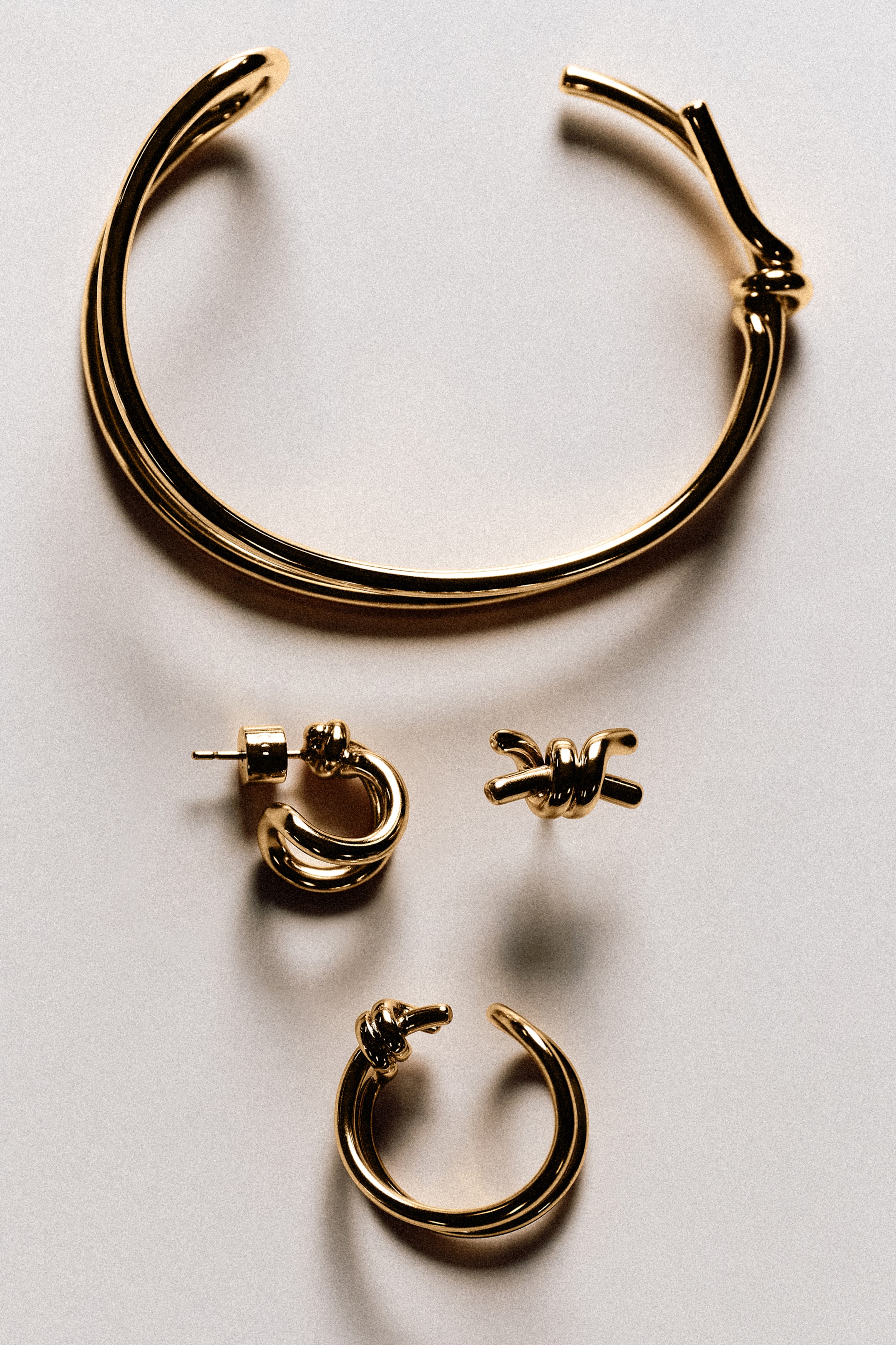 Größeres Bild ansehen: Ein offenes Armreif-Armband in Gold, ein kleiner Creolenohrring, ein knotenförmiger Ohrstecker und ein offener Ear Cuff werden präsentiert, alle mit Knotendetails. Das Armreif-Armband zeigt ein zweireihiges Design und einen Knoten an einem Ende, während der Creolenohrring und der Ear Cuff Knotendetails aufweisen und der Ohrstecker eine Knotenform hat.