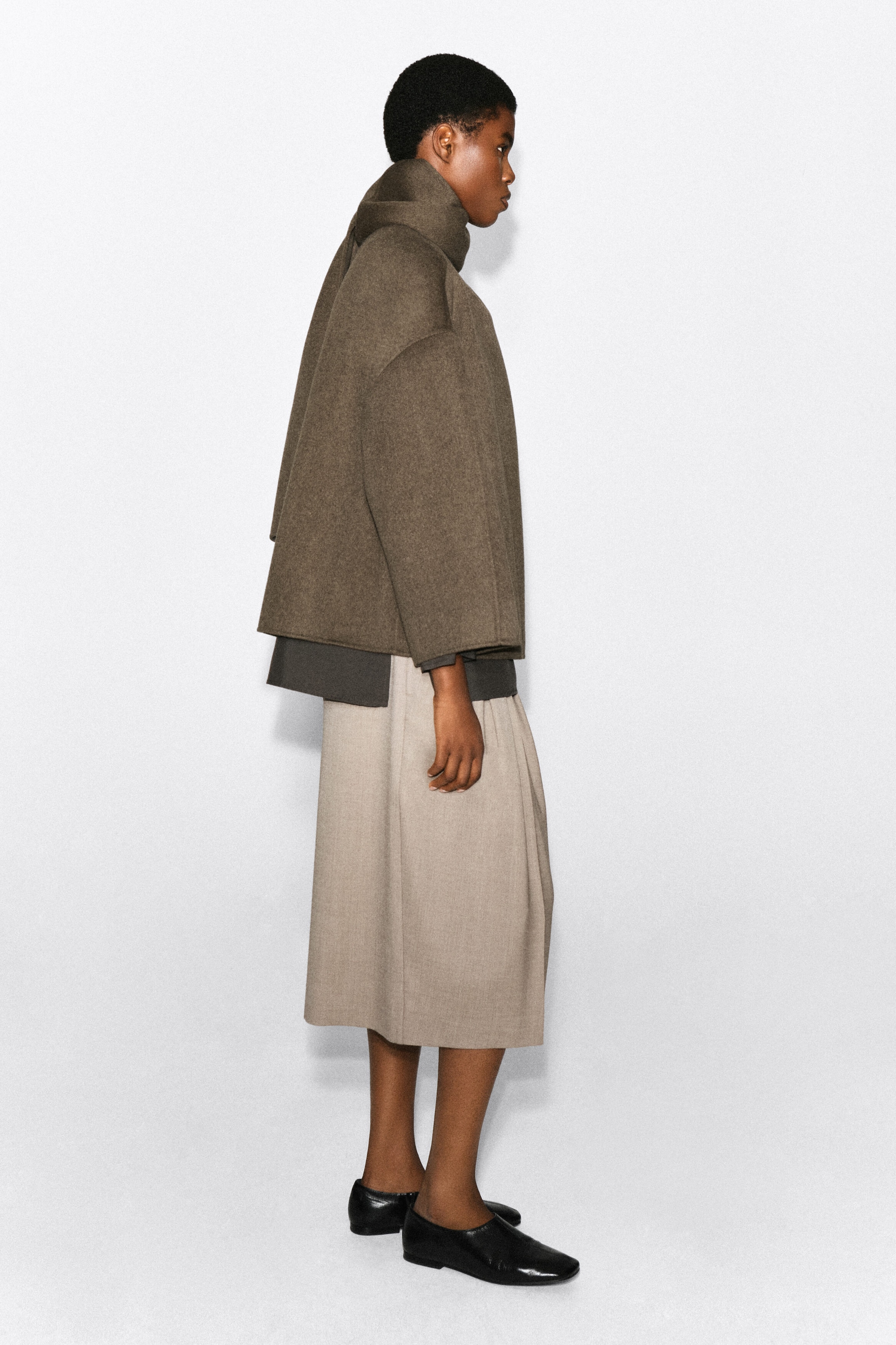 A double-faced wool-blend scarf jacket in a textured brown hue is being worn by the model, shown from a side-profile view. The jacket features an integrated scarf collar draped around the neck, wide cropped sleeves, and a boxy silhouette layered over a dark grey top and beige midi skirt. A double-faced wool-blend scarf jacket in a textured brown hue is being worn by the model, shown from a side-profile view. The jacket features an integrated scarf collar draped around the neck, wide cropped sleeves, and a boxy silhouette layered over a dark grey top and beige midi skirt.