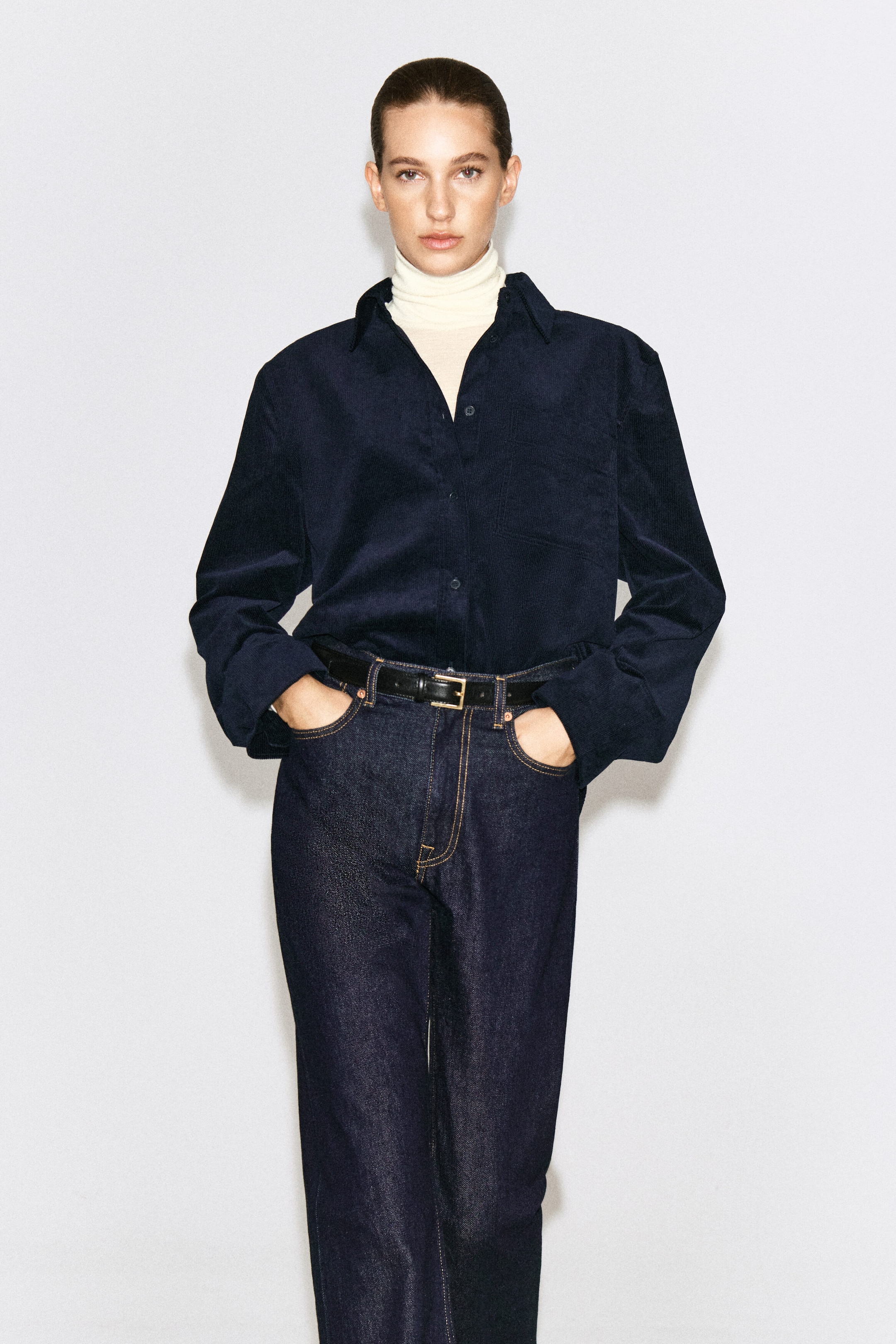 The model is wearing a dark navy cotton corduroy shirt layered over a cream turtleneck and tucked into dark wash jeans with a black belt. The long-sleeve shirt features a classic collar and a button-front closure. The model is wearing a dark navy cotton corduroy shirt layered over a cream turtleneck and tucked into dark wash jeans with a black belt. The long-sleeve shirt features a classic collar and a button-front closure.