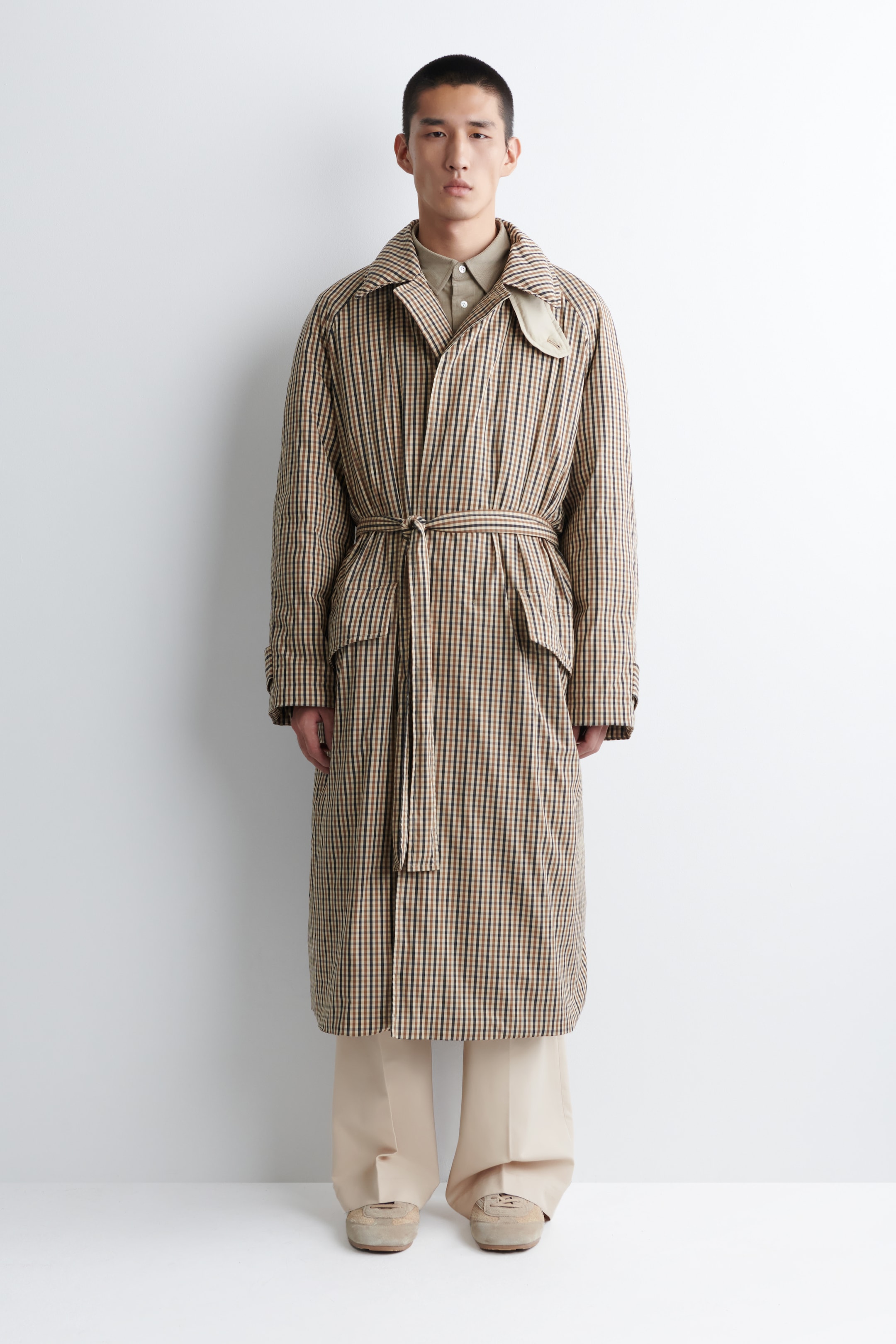 Grösseres Bild ansehen: Ein langer, vorn offener Mantel in einem braunen, schwarzen und beigen Gingham-Muster weist eine entspannte Passform, einen Umlegekragen, Raglanärmel und eine Sturmpatte auf der rechten Schulter auf. Zwei große aufgesetzte Taschen sind vorne sichtbar, mit einem Gürtel aus dem gleichen Stoff, der die Taille betont, und er wird über einem hellbraunen Hemd mit Kragen getragen.