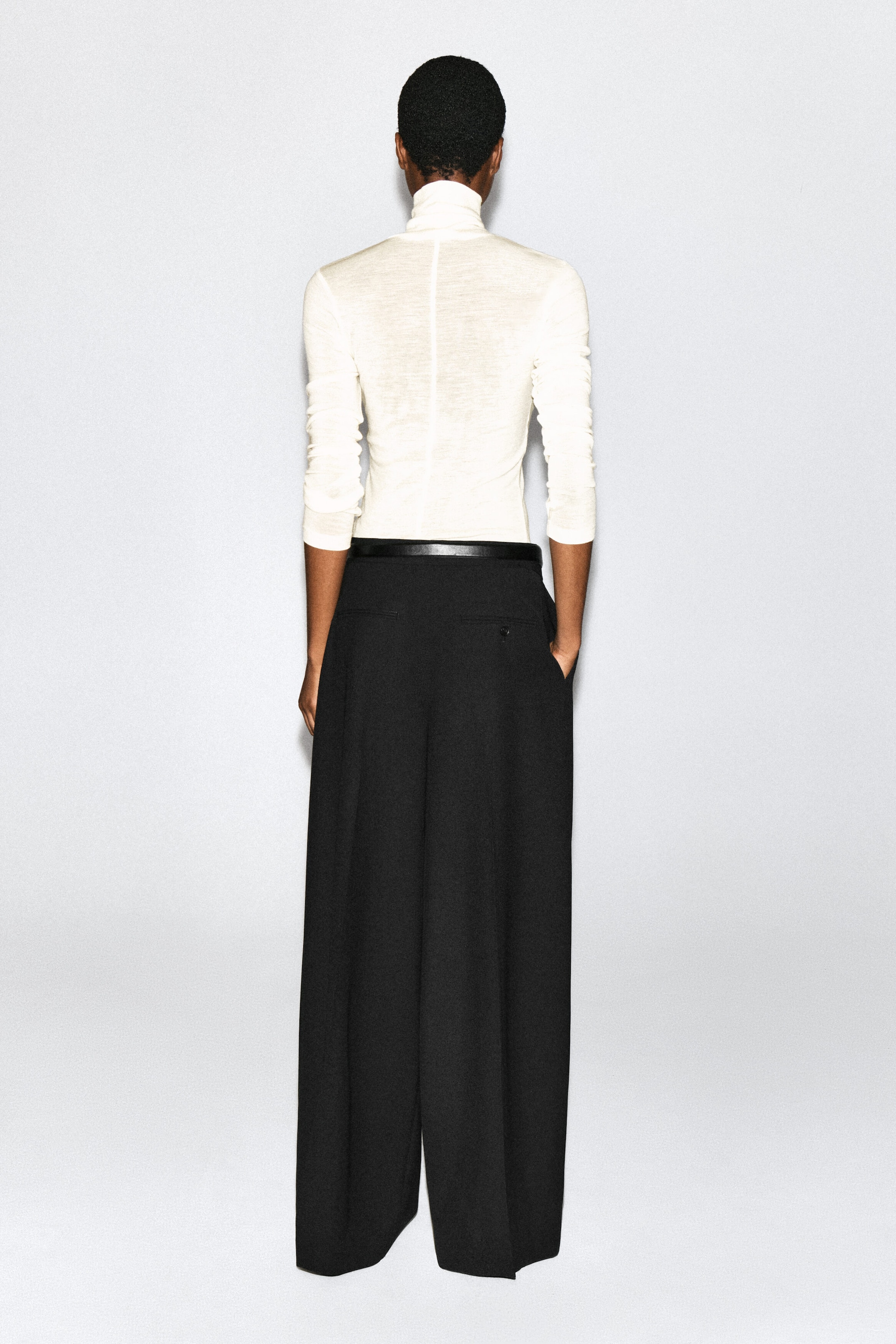 The model is wearing black pleated wide-leg pants, shown from the back, styled with a thin black belt and a tucked-in cream turtleneck top. The model is standing with one hand resting in the pants' side pocket. The model is wearing black pleated wide-leg pants, shown from the back, styled with a thin black belt and a tucked-in cream turtleneck top. The model is standing with one hand resting in the pants' side pocket.