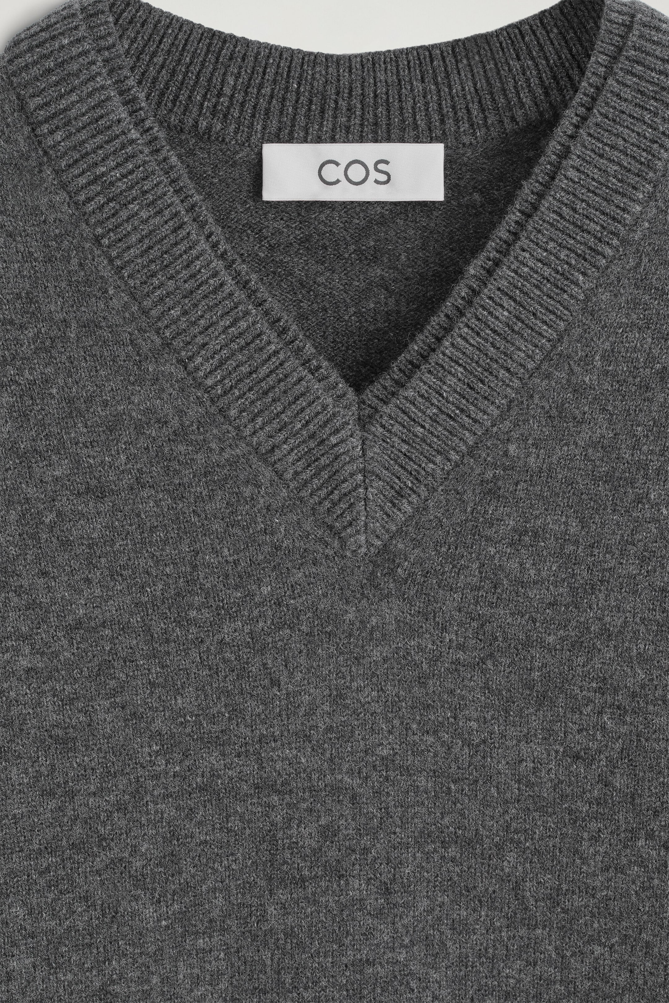 The Merino Wool Layered V-Neck Jumper is shown in a close-up front view, featuring a dark grey knit texture. A ribbed V-neckline layers over an inner ribbed crew neck, which displays a white "COS" brand label sewn on the inside. The Merino Wool Layered V-Neck Jumper is shown in a close-up front view, featuring a dark grey knit texture. A ribbed V-neckline layers over an inner ribbed crew neck, which displays a white "COS" brand label sewn on the inside.