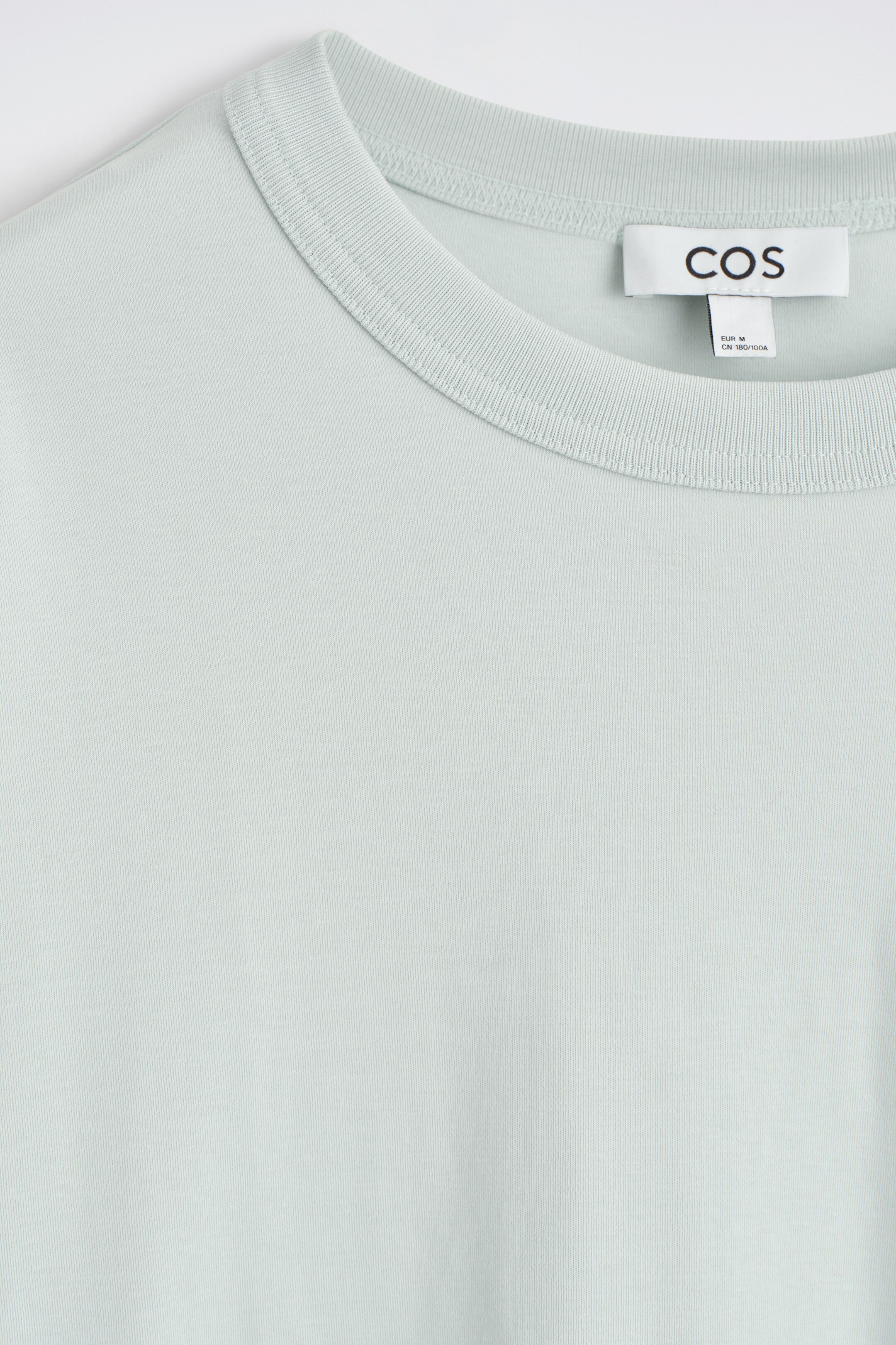View larger image: A light green t-shirt is being presented from the FRONT, showcasing its ribbed crew neck and smooth fabric. A white brand tag with "COS" is visible on the inside back of the neckline.