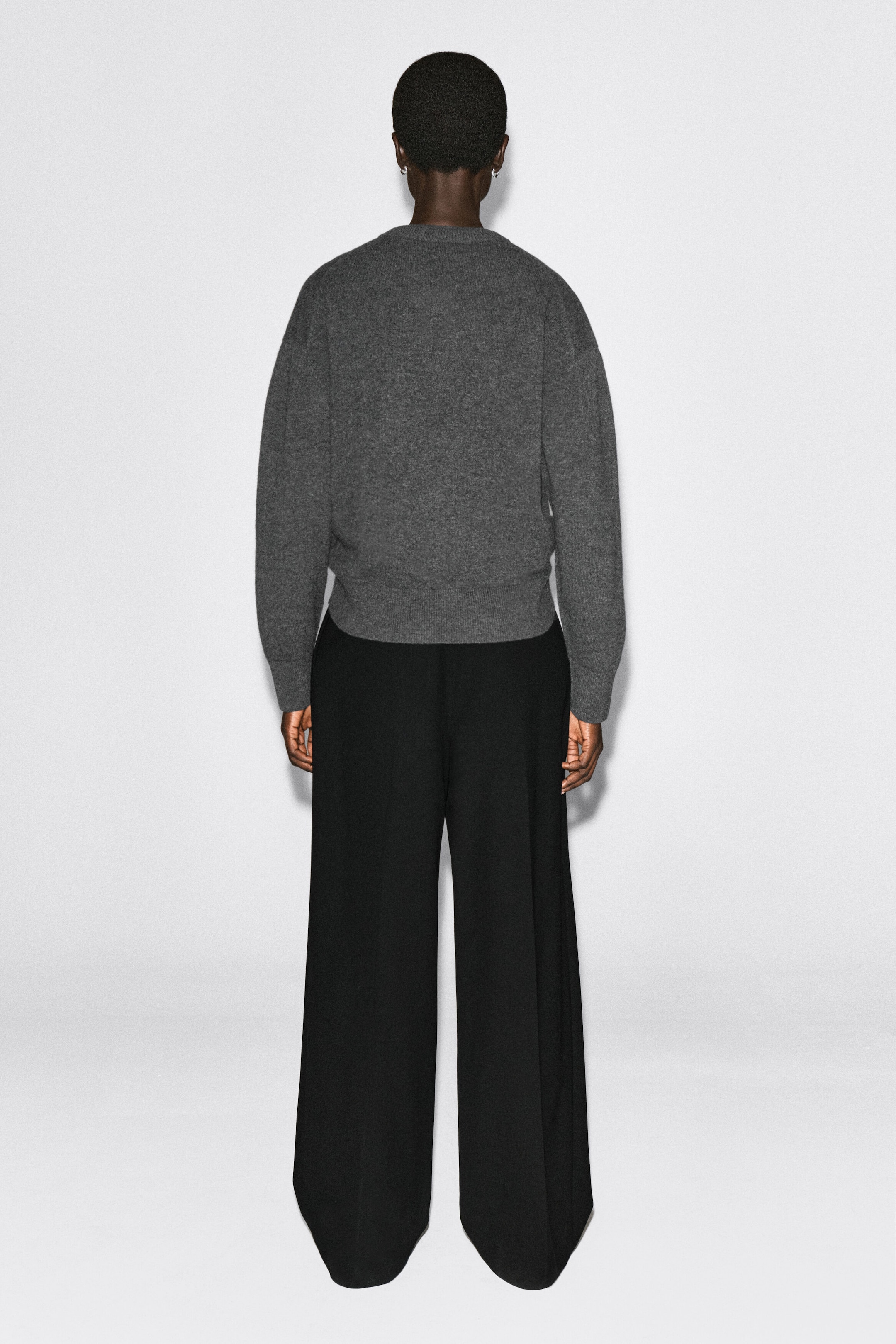 The merino wool sweater is shown from the back, featuring a relaxed fit with dropped shoulders, ribbed cuffs, and a ribbed hem. The charcoal gray knit sits at the waist and is styled with black wide-leg trousers. The merino wool sweater is shown from the back, featuring a relaxed fit with dropped shoulders, ribbed cuffs, and a ribbed hem. The charcoal gray knit sits at the waist and is styled with black wide-leg trousers.