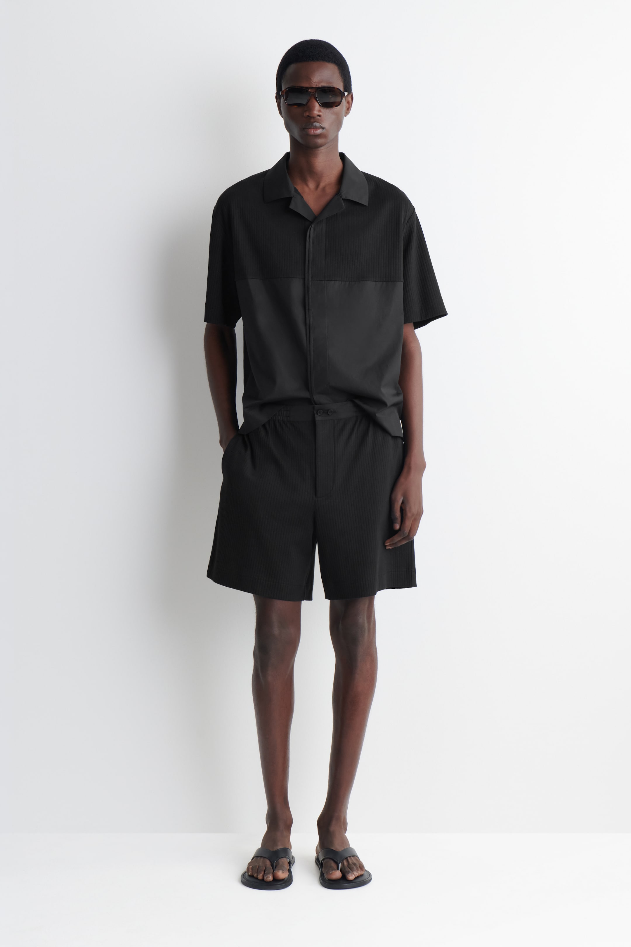 Größeres Bild ansehen: Schwarze gerippte Baumwoll-Jersey-Shorts mit einem kontrastierenden Baumwoll-Popeline-Bund werden vom Model getragen. Diese Shorts im lässigen Schnitt verfügen über sichtbare Seitentaschen und einen Knopfverschluss und werden mit einem passenden schwarzen Hemd kombiniert.