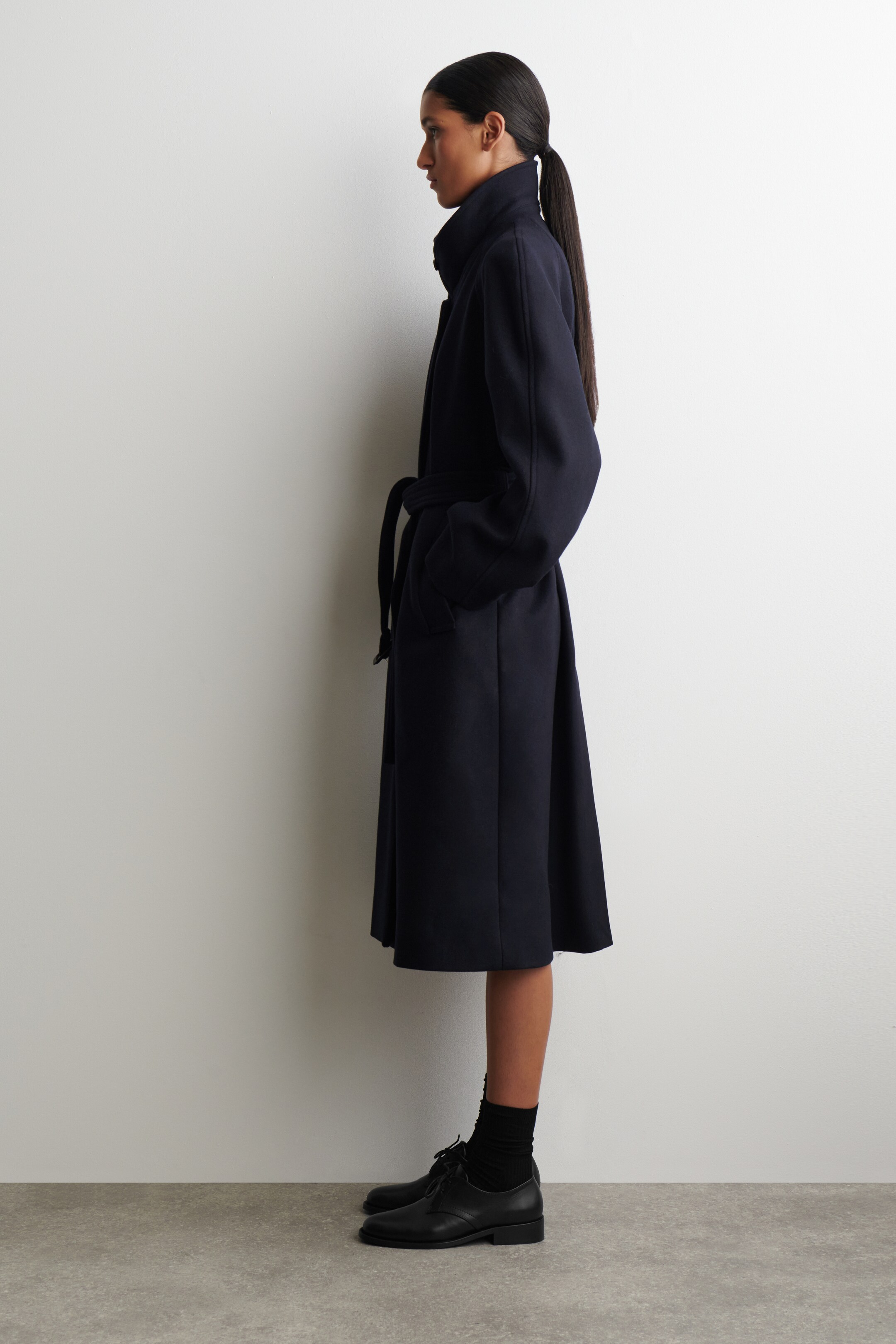 Ingrandisci l'immagine: Un cappotto blu scuro è presentato di profilo, con un colletto alto, maniche lunghe e mani nelle tasche laterali. Il cappotto è stretto in vita con una cintura dello stesso tessuto, e la sua silhouette a trapezio si allarga verso un orlo a lunghezza midi, con scarpe oxford nere e calzini visibili sotto.