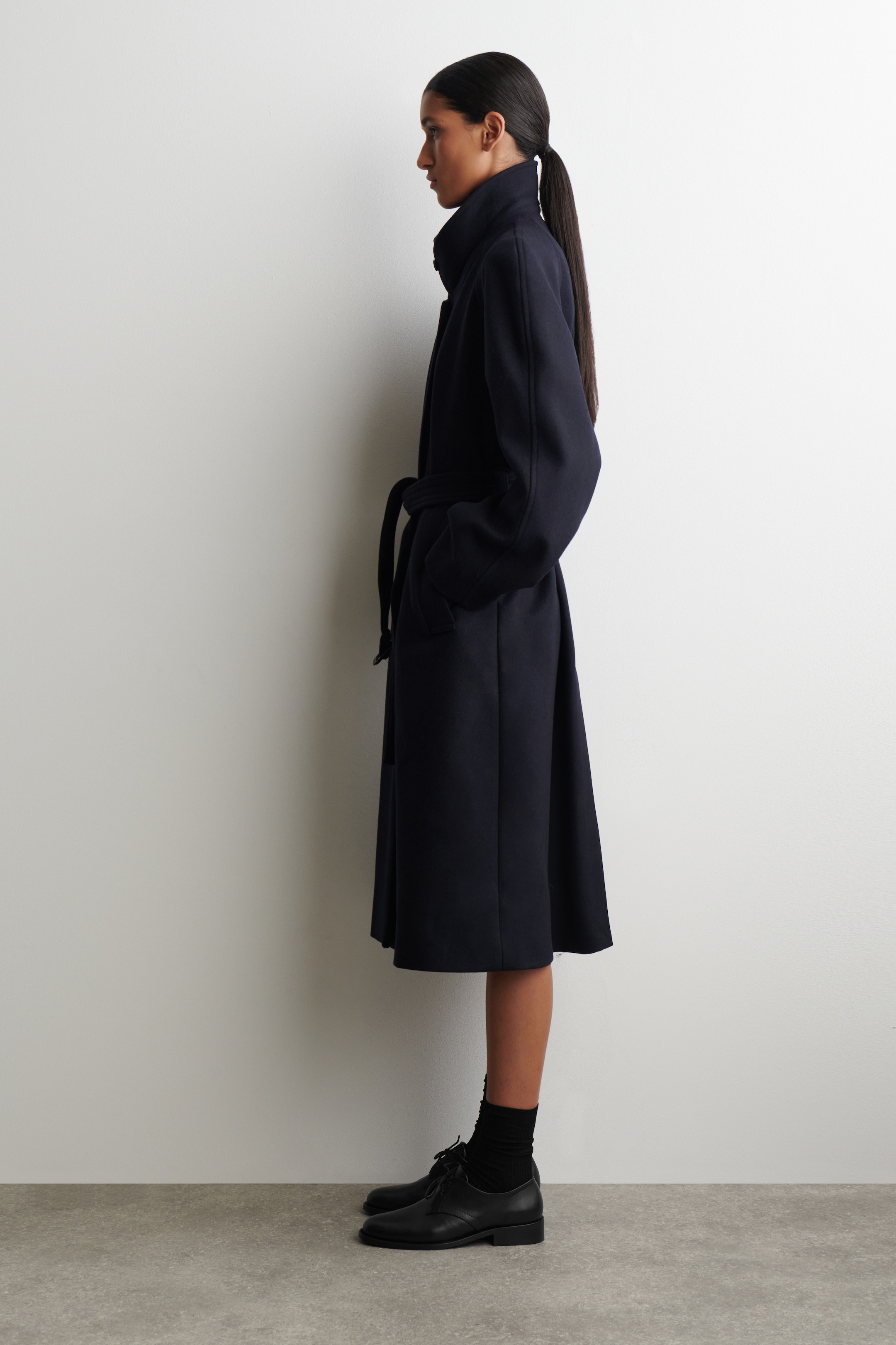 Agrandir l'image: Un manteau bleu marine foncé est présenté de profil, avec un col montant, des manches longues et des mains reposant dans les poches latérales. Le manteau est ceinturé à la taille avec une ceinture en tissu assorti, et sa silhouette en ligne A s'évase vers un ourlet mi-longueur, avec des chaussures oxford noires et des chaussettes visibles en dessous.