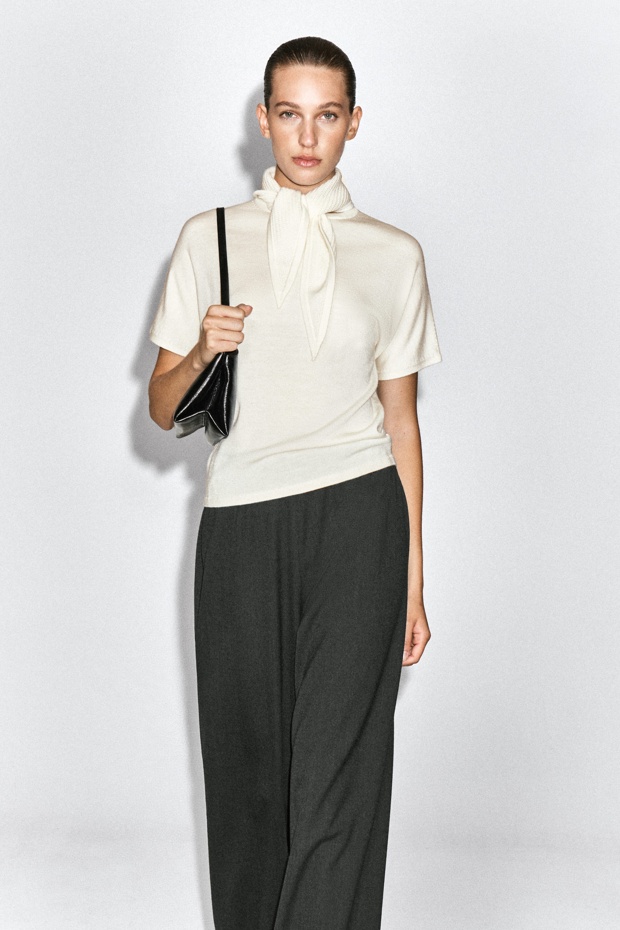 A scarf-detail merino wool-blend T-shirt in cream is worn by the model, featuring short sleeves and an integrated scarf tied at the neck. The T-shirt is styled with dark wide-leg trousers, and a black leather shoulder bag is carried.