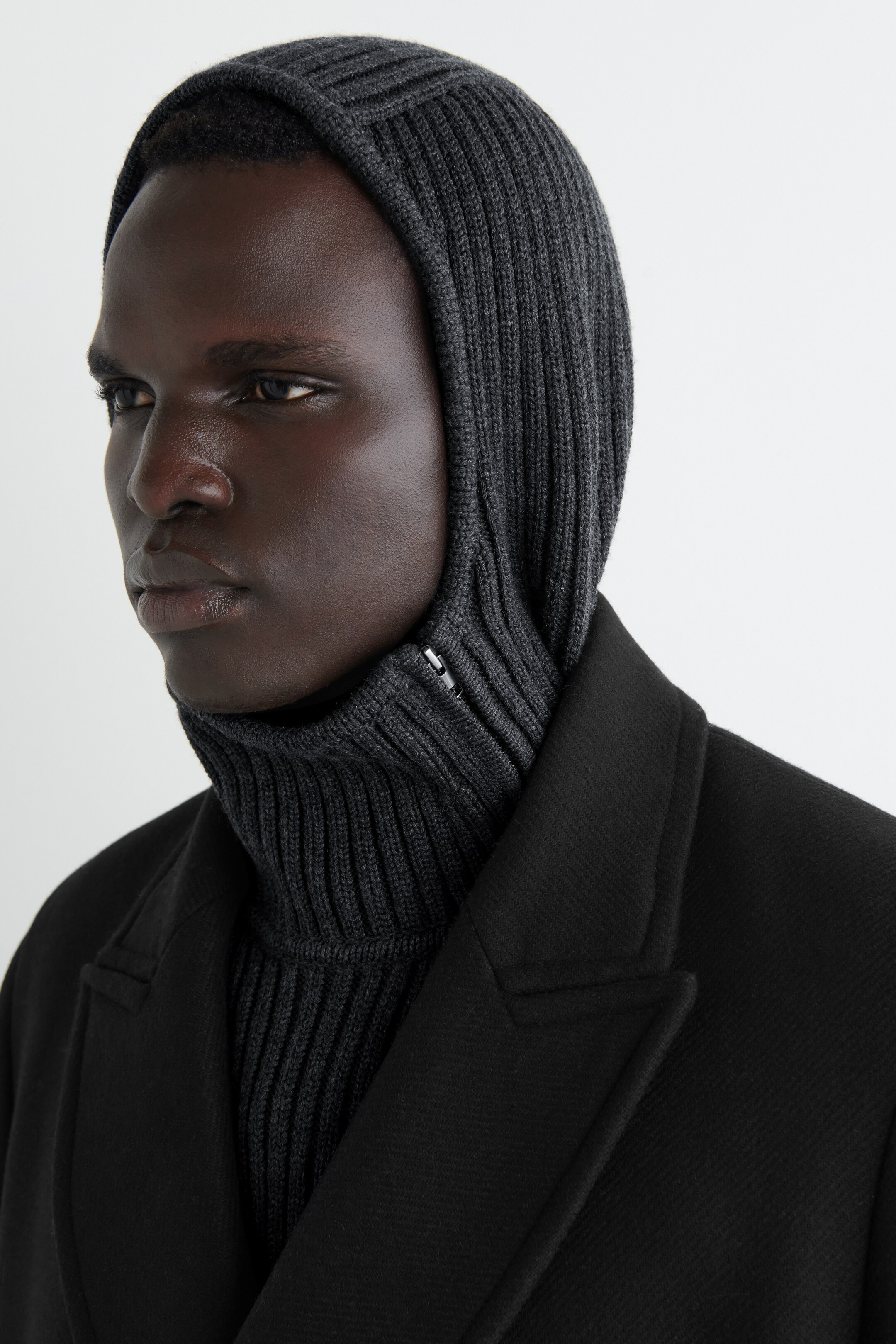 Agrandir l'image: Un hoodie mélangé en charbon, présentant une construction en tricot à larges côtes, est porté avec sa capuche tubulaire couvrant la tête et le cou. Une fermeture éclair est visible sur la zone de l'épaule gauche du hoodie, qui est superposé sous un manteau noir.