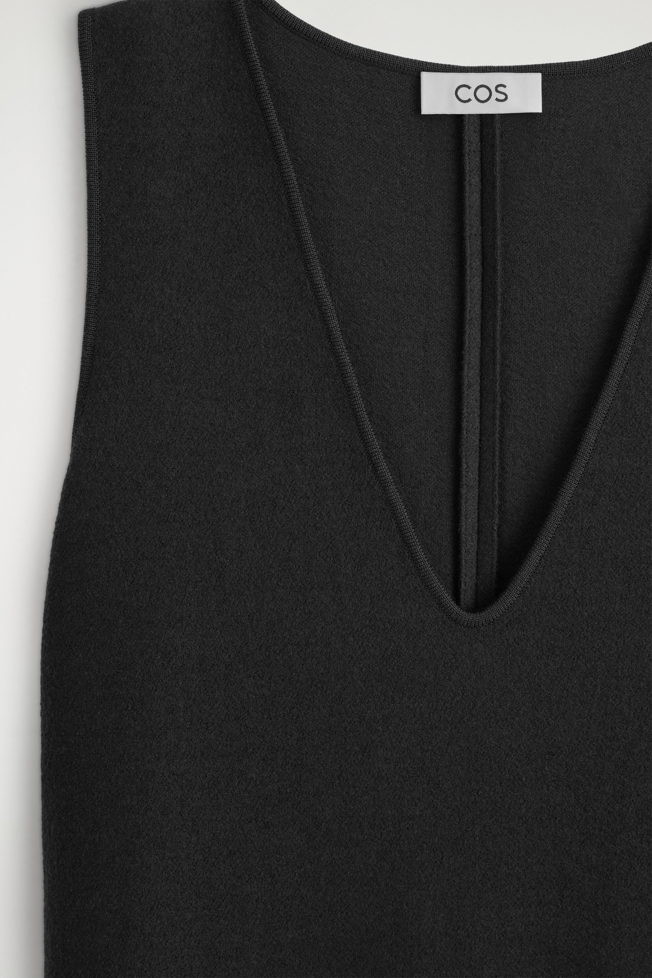 The boiled wool-blend V-neck midi dress is shown in a close-up front view, featuring a deep V-neckline with fine ribbed trim. The sleeveless design highlights a textured, black felt-like fabric, with a white brand label visible at the back collar. The boiled wool-blend V-neck midi dress is shown in a close-up front view, featuring a deep V-neckline with fine ribbed trim. The sleeveless design highlights a textured, black felt-like fabric, with a white brand label visible at the back collar.