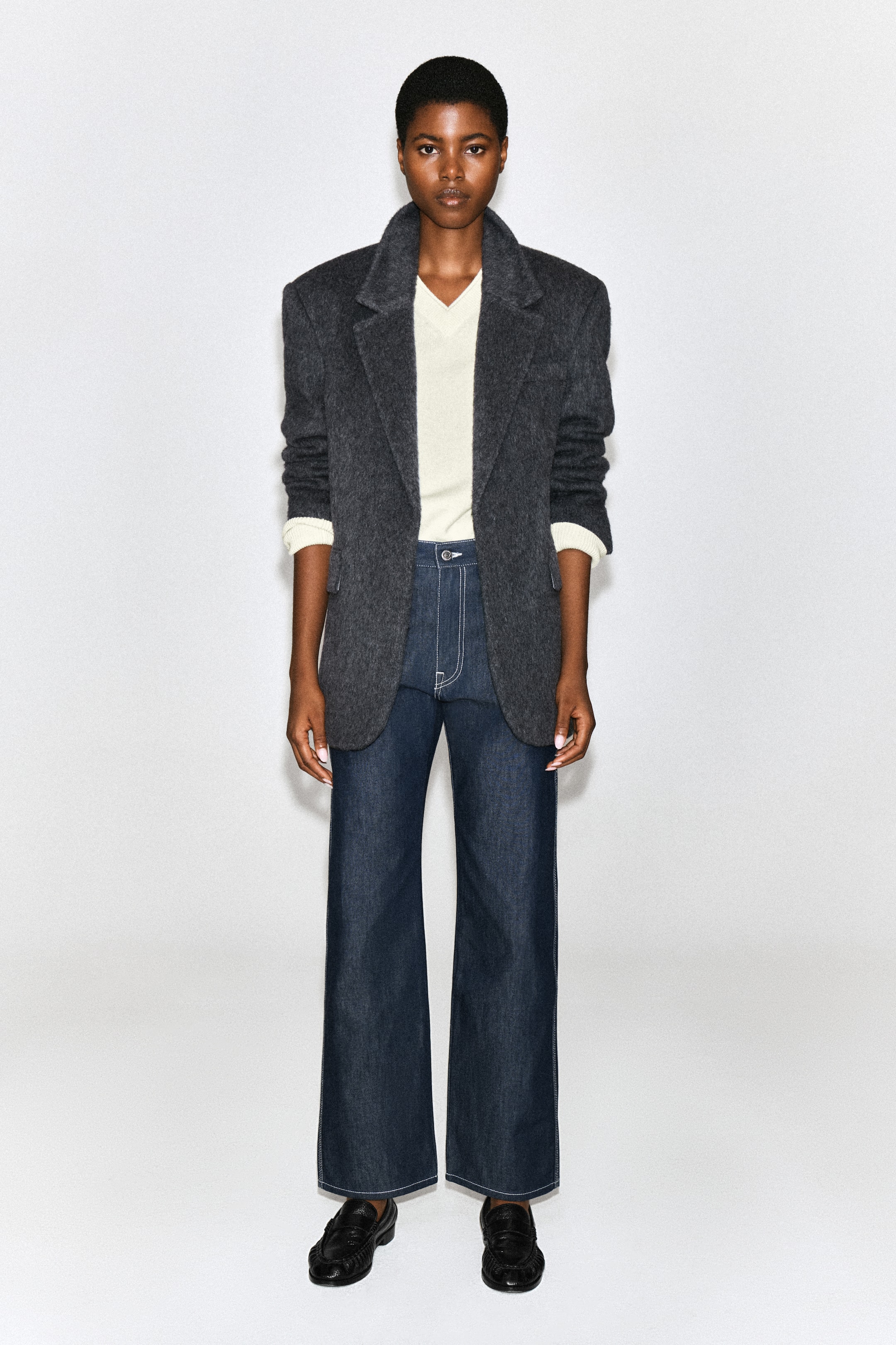 Ver imagen más grande: Los vaqueros rectos CHORD de denim azul oscuro con pespuntes vistos los lleva la modelo, con un corte de tiro alto y un marcado pespunte blanco en contraste. Los vaqueros se combinan con un jersey crema de cuello en V por dentro, un blazer oversize gris oscuro y unos mocasines negros.