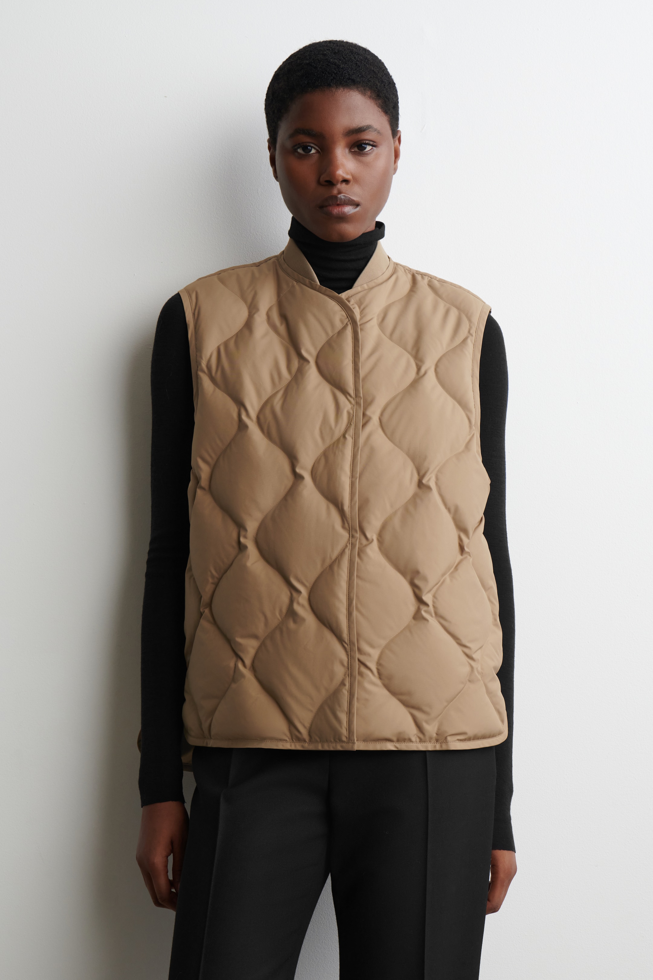 View larger image: A tan quilted vest is being worn front-facing, featuring a band collar, a front snap placket, and a diamond-shaped quilting pattern, layered over a black turtleneck.