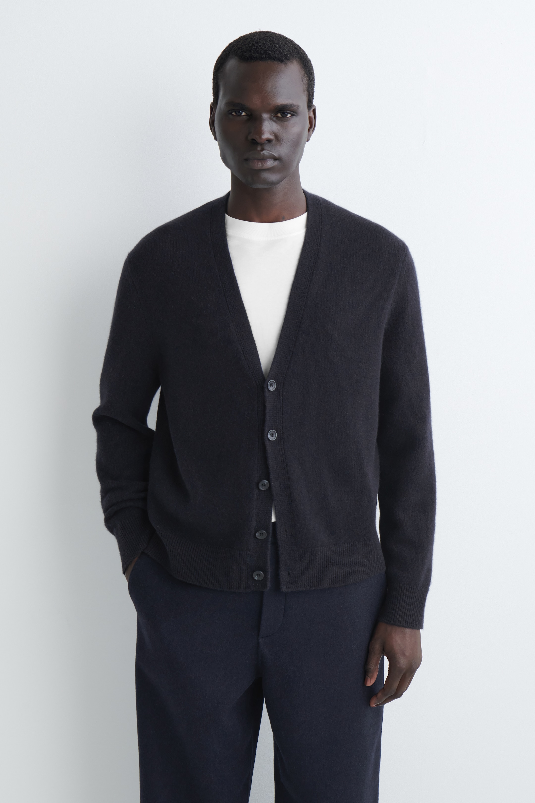 Größeres Bild ansehen: Eine dunkelblaue Strickjacke wird frontal präsentiert und liegt über einem weißen T-Shirt. Sie verfügt über einen V-Ausschnitt, einen Verschluss mit sechs Knöpfen vorne, lange Ärmel mit gerippten Bündchen und einen gerippten Saum.