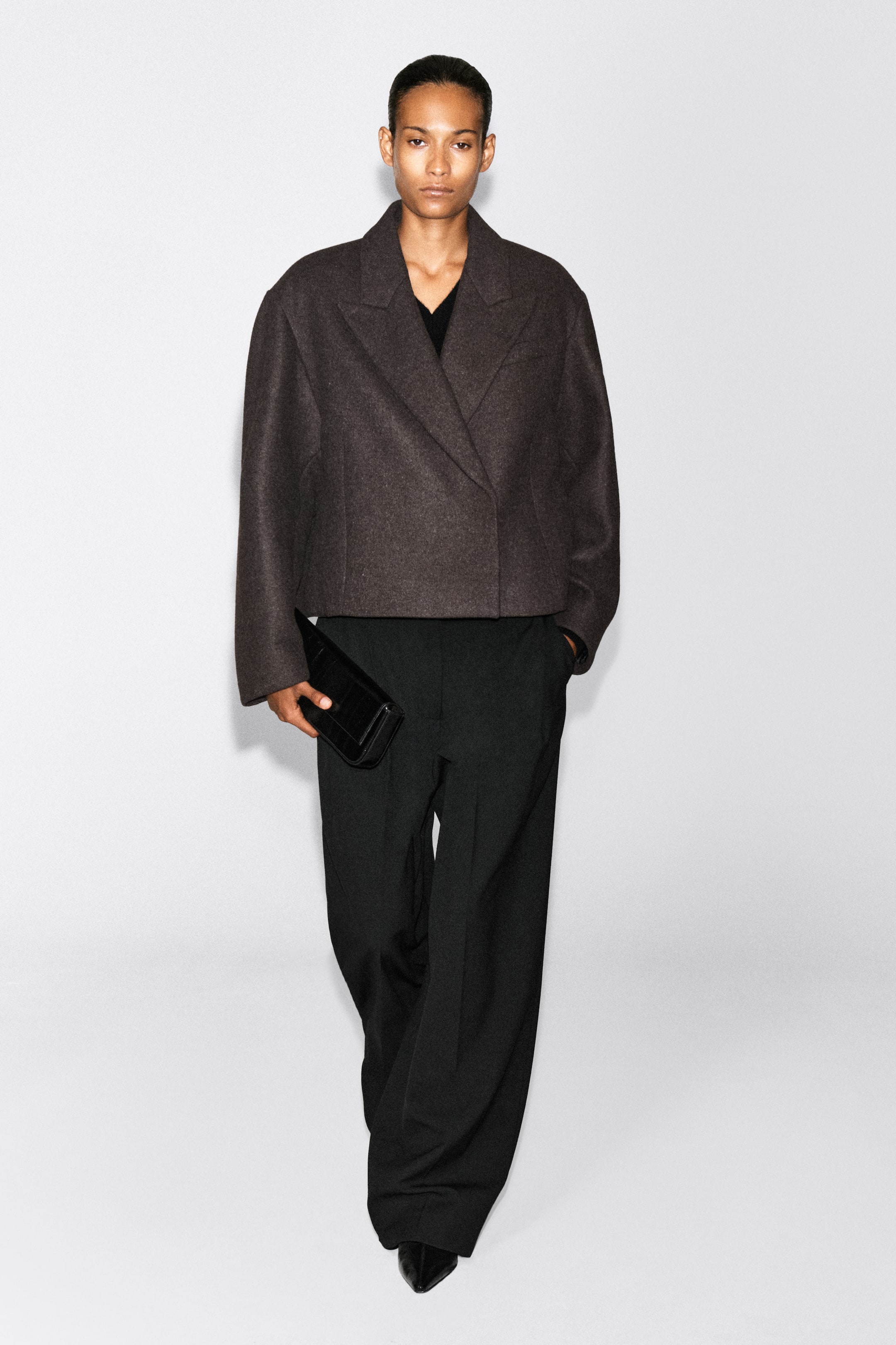 Ver imagem maior: Um blazer oversize cropped em mistura de lã fervida, em castanho-escuro, é usado pela modelo, apresentando um corte boxy, ombros descaídos e lapelas largas com entalhe. O blazer é combinado com calças pretas de perna larga, enquanto a modelo segura uma pequena clutch preta.