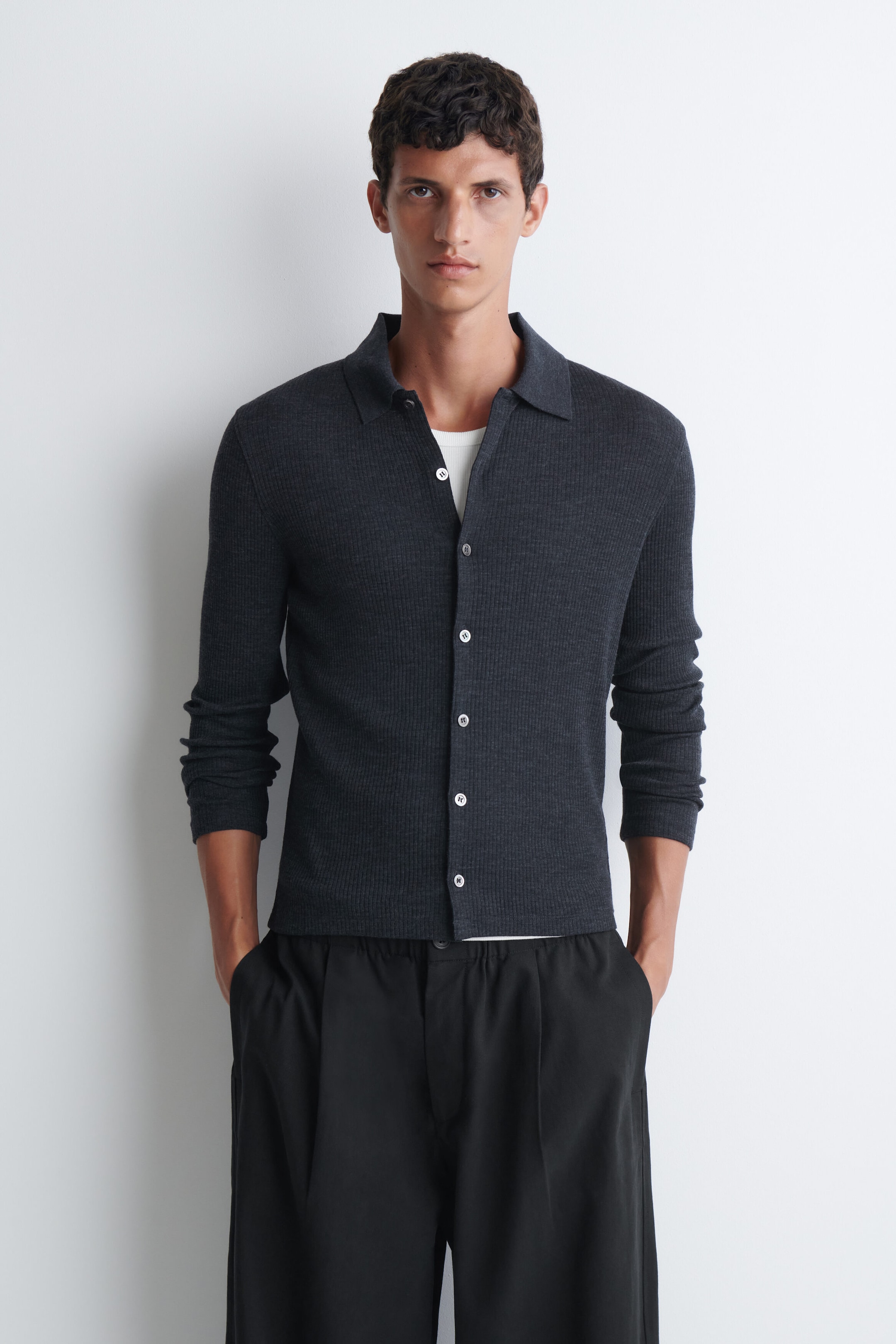 A dark grey, ribbed-knit long-sleeve polo shirt with a button-front placket is being worn by the model over a white T-shirt. The polo shirt is styled with the top buttons undone and paired with black trousers.