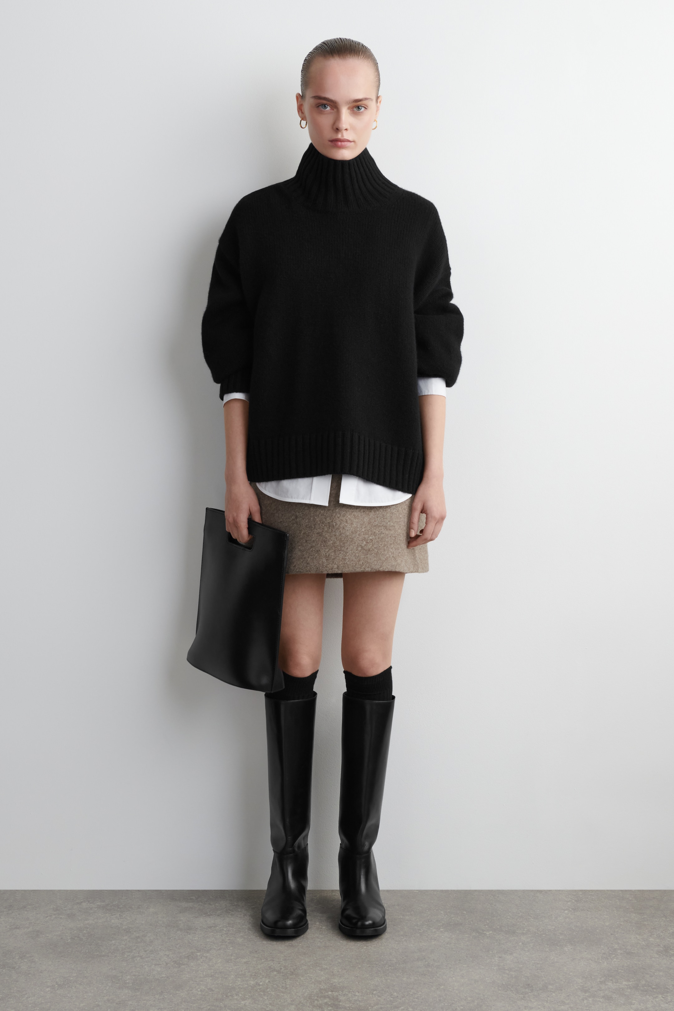 Visualizza immagine più grande: Un maglione dolcevita nero è presentato frontalmente, con un alto colletto a coste, maniche lunghe con polsini a coste e un orlo a coste. Viene stratificato su una camicia bianca col colletto, con i polsini e l'orlo della camicia che si estendono oltre il maglione.