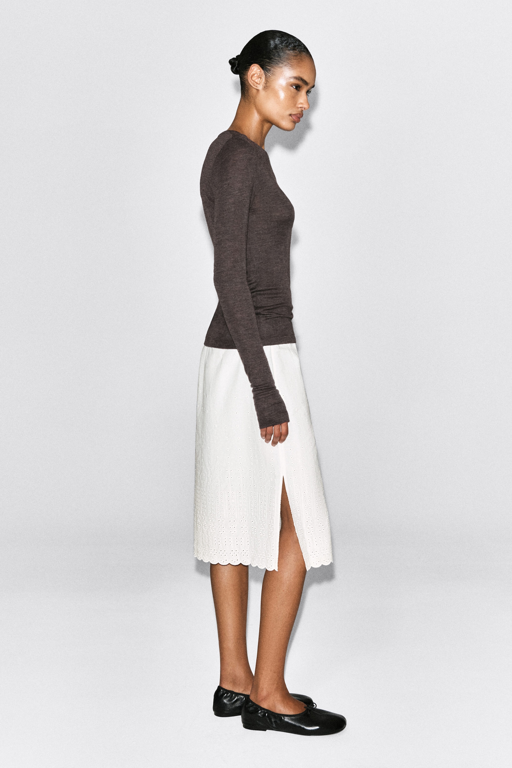 A slim merino wool top in dark brown is shown from a right-facing profile view, highlighting its form-fitting silhouette and long sleeves with extended cuffs. The lightweight, crew-neck top is worn untucked over a white skirt. A slim merino wool top in dark brown is shown from a right-facing profile view, highlighting its form-fitting silhouette and long sleeves with extended cuffs. The lightweight, crew-neck top is worn untucked over a white skirt.