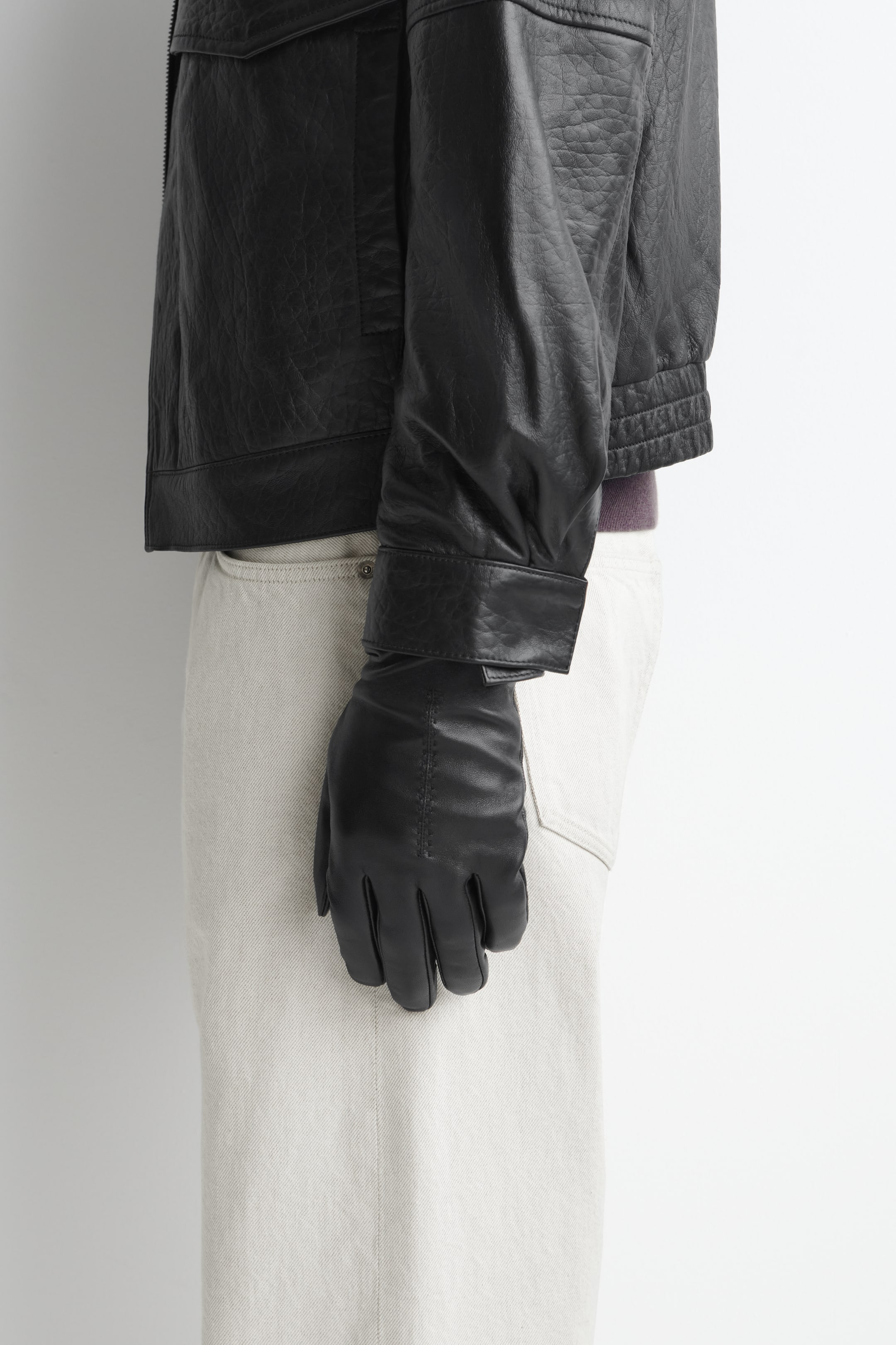 Größeres Bild ansehen: Ein schwarzer Lederhandschuh wird an einer nach links gerichteten Hand getragen und zeigt eine glatte Textur mit genähten Pintuck-Details auf dem Handrücken. Der Handschuh ist stilvoll unter dem Ärmelaufschlag einer strukturierten schwarzen Lederjacke verborgen.