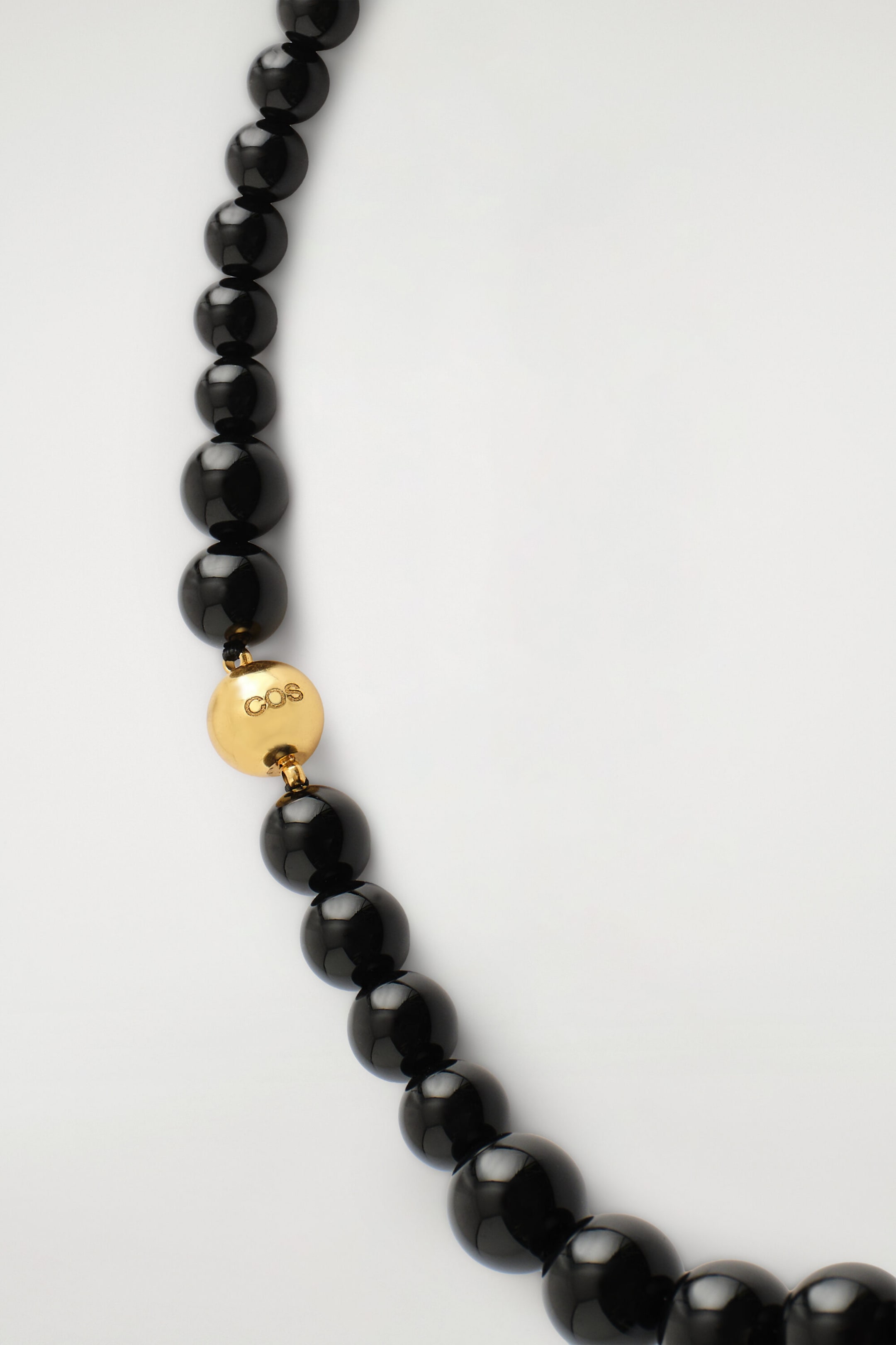 Ce collier de perles met en valeur des perles noires irrégulières et brillantes, dont la taille augmente progressivement. Un fermoir sphérique à fini doré poli, gravé « COS », assure la fermeture.