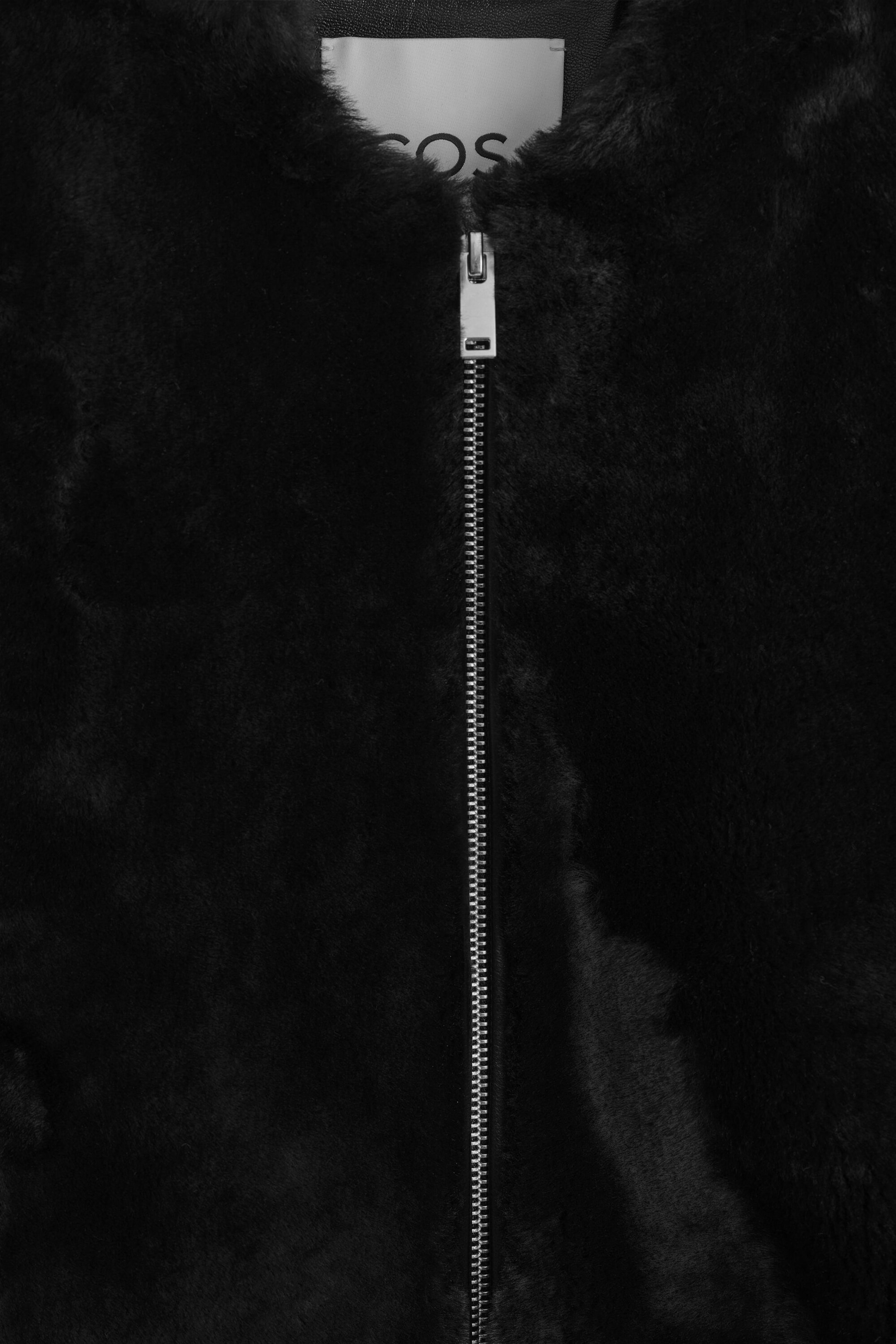 The black padded shearling bomber jacket is shown in a close-up front view, highlighting its plush, textured shearling surface. The jacket features a central silver-tone metal zipper closure and a partially visible white label at the inner neckline.