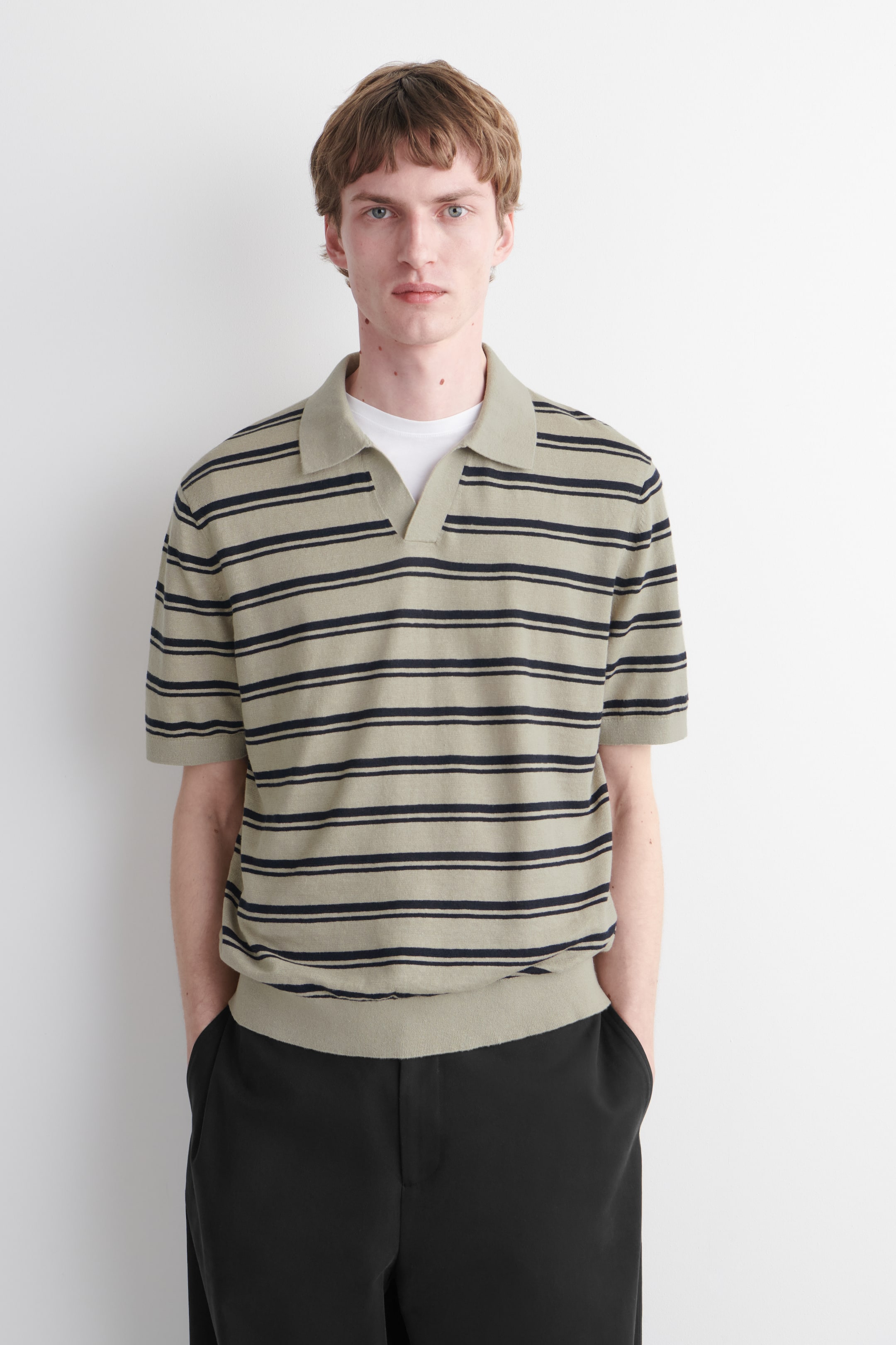 Grösseres Bild ansehen: Das Modell trägt ein gestricktes Poloshirt mit hellgrünen und dunklen horizontalen Streifen. Das Shirt hat einen offenen Kragen, kurze Ärmel und einen gerippten Saum und ist über einem weissen T-Shirt geschichtet.