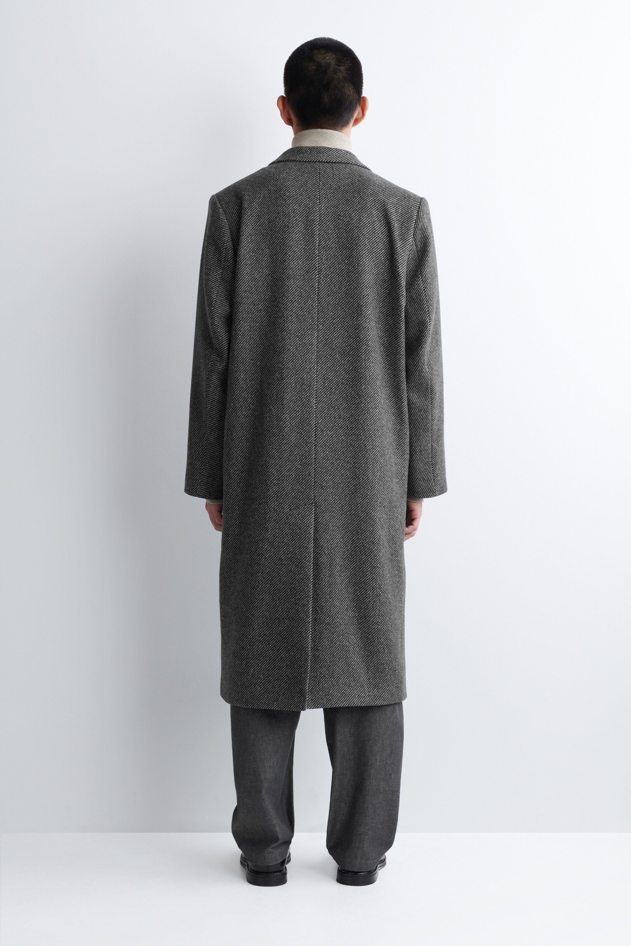 Agrandir l'image: Un long manteau gris foncé est présenté de dos, présentant une coupe décontractée et un tissu richement texturé en laine recyclée. Il est porté par-dessus un col roulé de couleur claire.