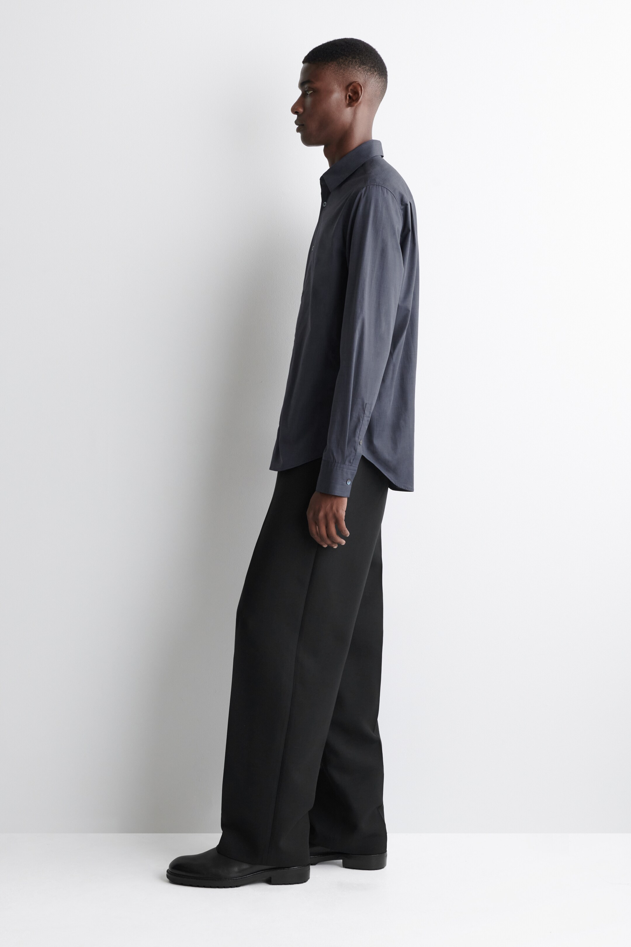 Agrandir l'image: Une chemise gris foncé à col, avec des manches longues et des poignets boutonnés, est présentée de côté, mettant en valeur une coupe décontractée. Elle est associée à un pantalon noir à jambes larges.
