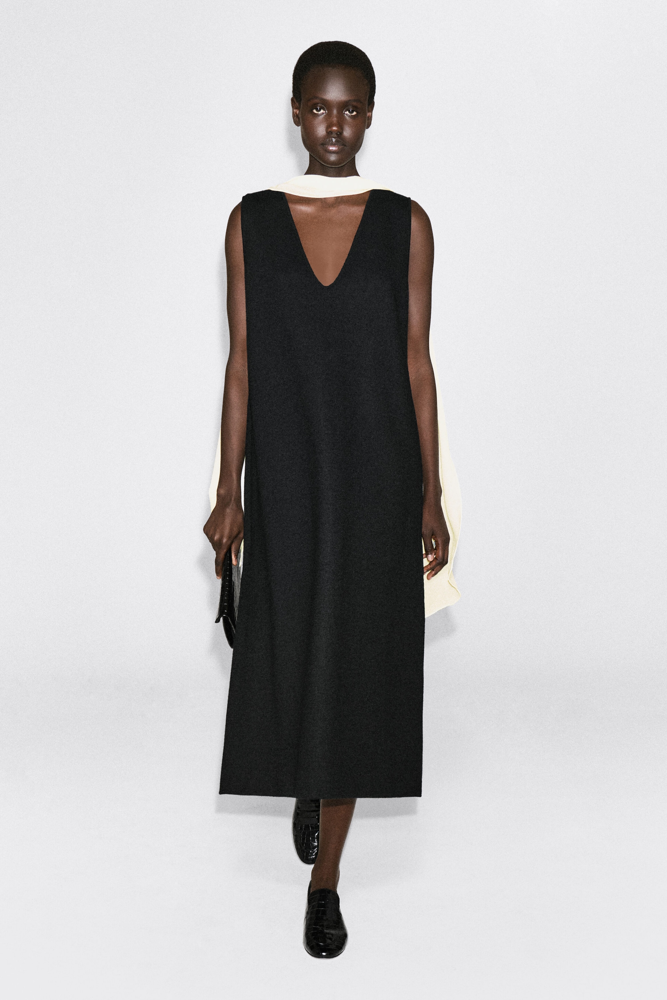 A black boiled wool-blend V-neck midi dress is shown from the front, featuring a sleeveless design, a deep V-neckline, and a relaxed, straight silhouette that falls to a midi length. The dress is styled with a contrasting cream-colored scarf draped around the neck. A black boiled wool-blend V-neck midi dress is shown from the front, featuring a sleeveless design, a deep V-neckline, and a relaxed, straight silhouette that falls to a midi length. The dress is styled with a contrasting cream-colored scarf draped around the neck.