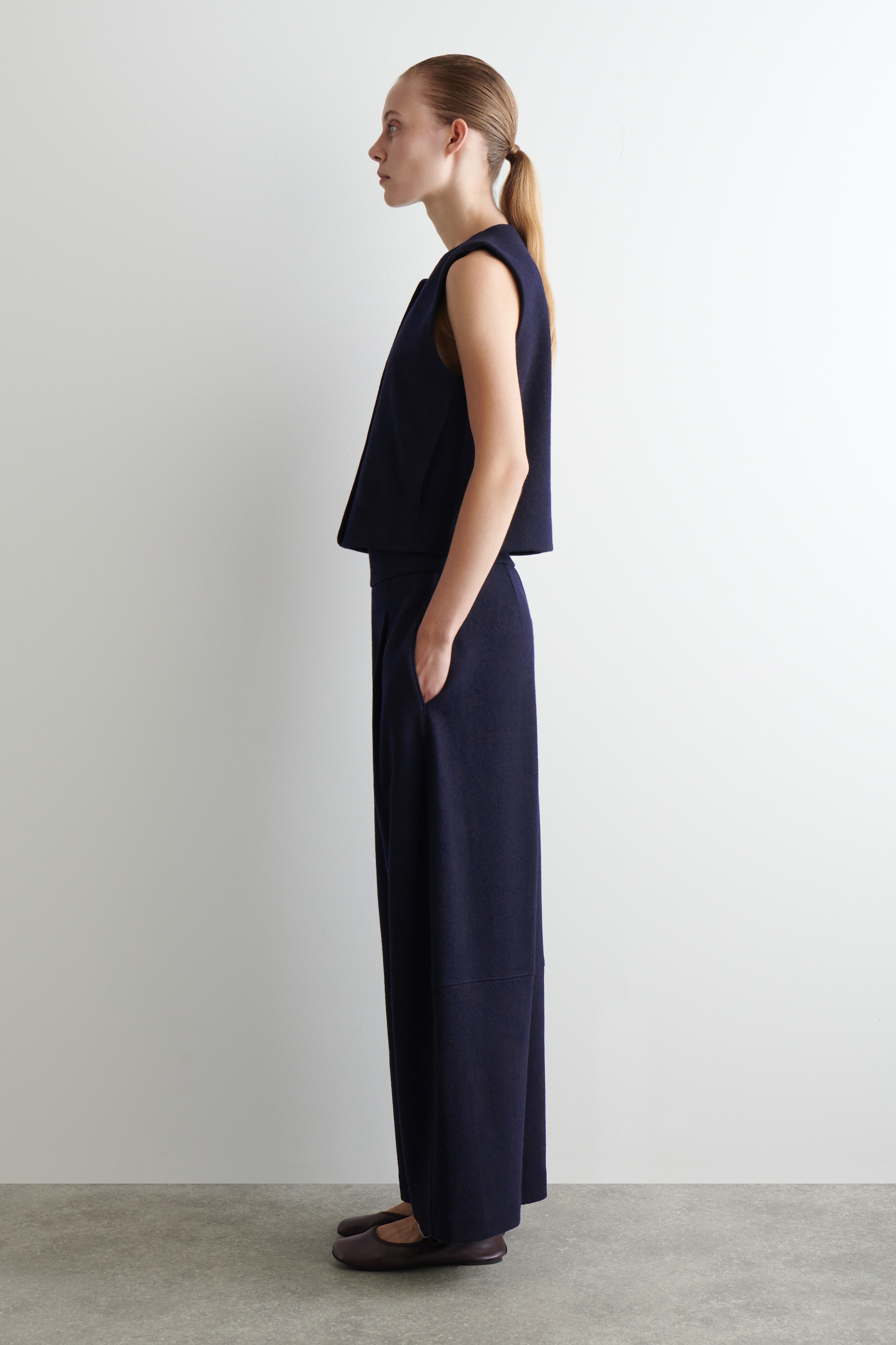 Ingrandisci l'immagine: Un completo blu navy a due pezzi viene presentato in vista laterale, mettendo in risalto la sua vestibilità rilassata e le linee pulite. L'outfit presenta un top corto senza maniche con un orlo dritto e pantaloni a gamba larga coordinati, che presentano tasche laterali e un sottile dettaglio di pannellatura orizzontale al ginocchio. Scarpe piatte e scure completano il look.