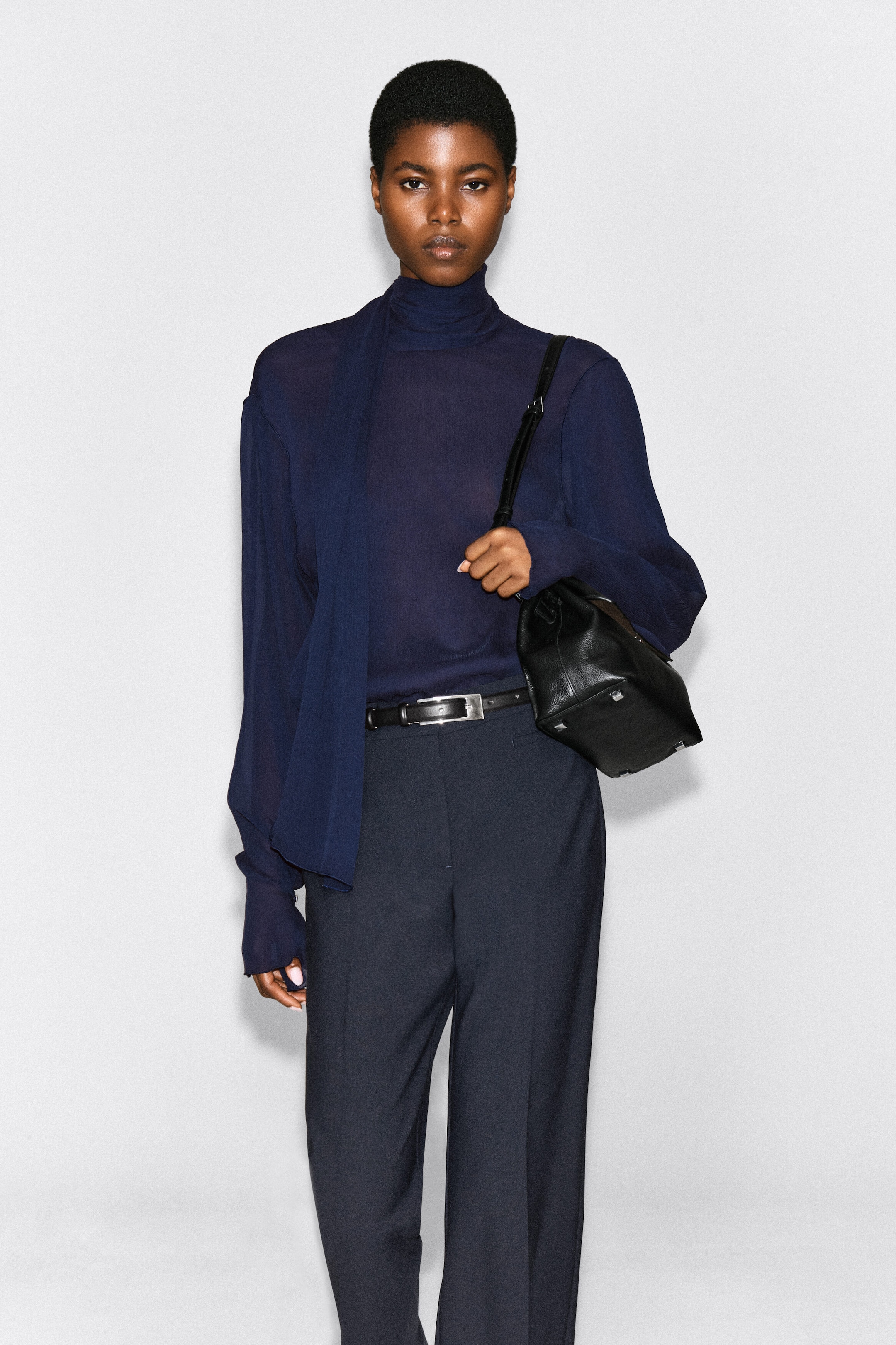 A dark navy blue, semi-sheer long-sleeved top featuring a high neck and an attached matching scarf draped over the shoulder is being worn by the model. The top is tucked into dark trousers with a black belt, and the model is carrying a black leather handbag over one shoulder. A dark navy blue, semi-sheer long-sleeved top featuring a high neck and an attached matching scarf draped over the shoulder is being worn by the model. The top is tucked into dark trousers with a black belt, and the model is carrying a black leather handbag over one shoulder.