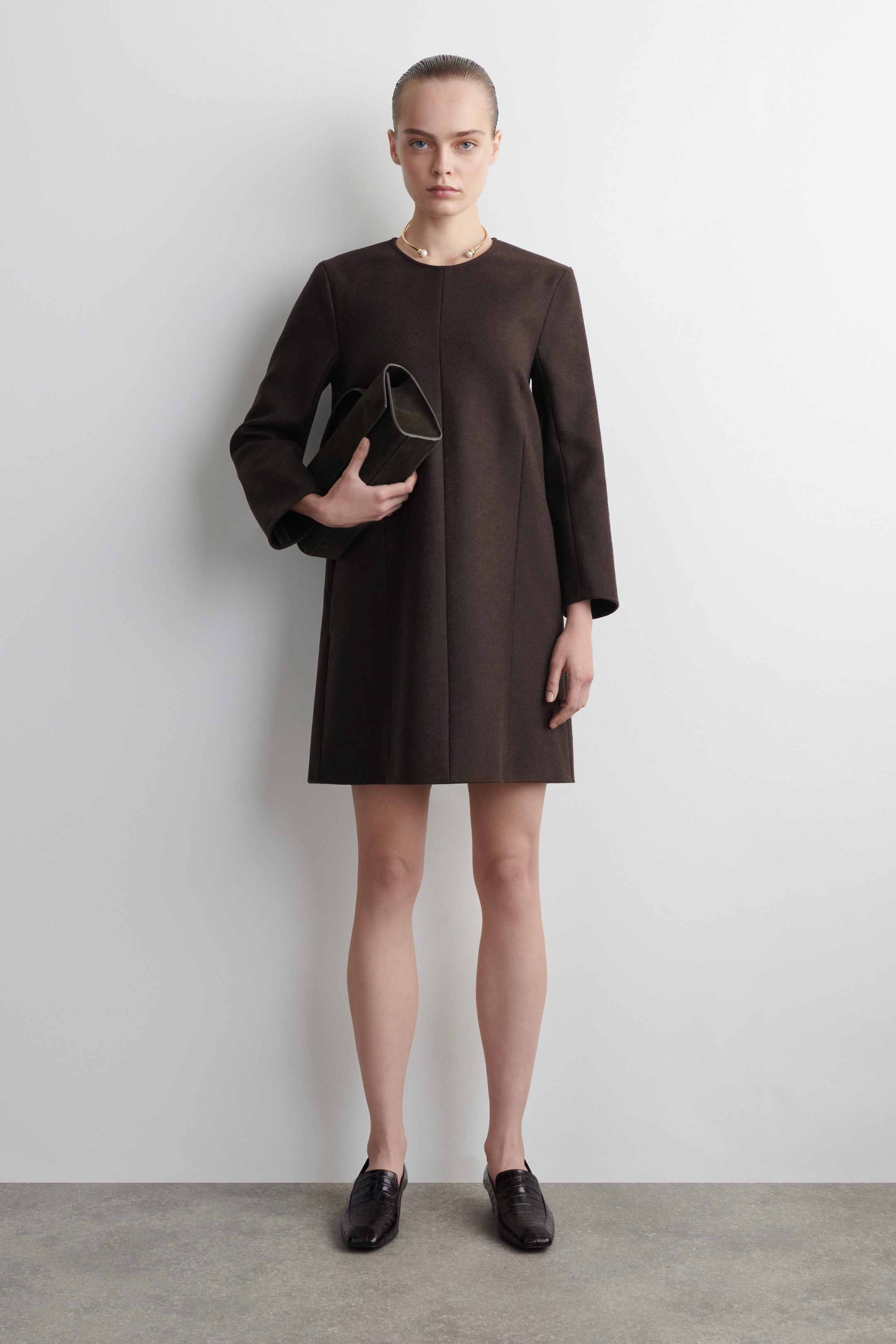 Ver imagen más grande: Un vestido mini en línea A de color marrón oscuro está siendo presentado en una vista frontal completa, con un escote tipo joya y mangas largas. El vestido muestra un ajuste relajado que se ensancha suavemente desde las caderas, con un collar dorado con dos perlas que descansan sobre su escote, y un bolso de mano marrón oscuro sostenido contra él en el lado izquierdo.