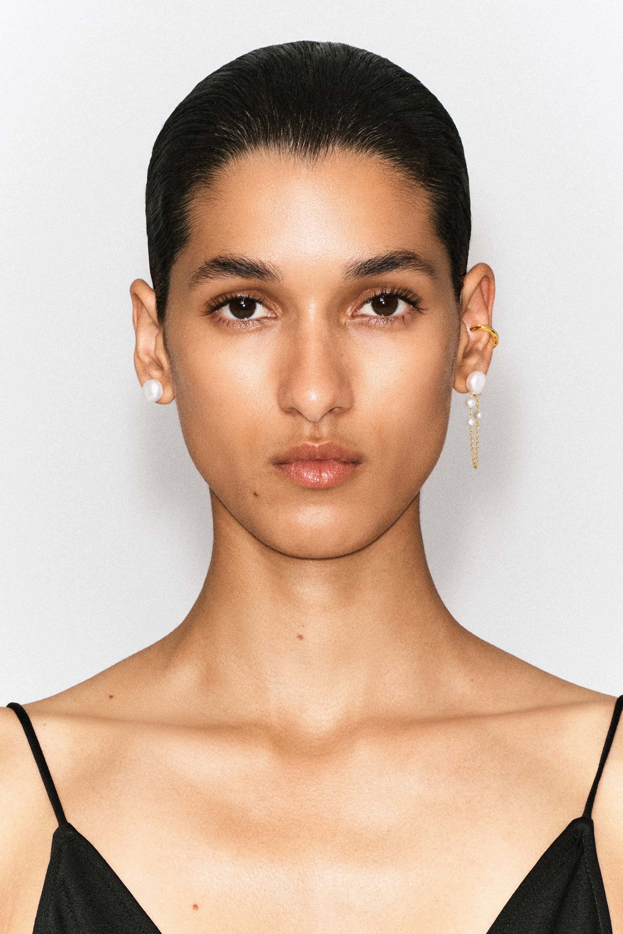 Grotere afbeelding bekijken: Een asymmetrische oorbellenset wordt van voren gestyled, met op één oorlel een enkele ronde witte parelstekker en op de andere oorlel een oorbel met hangertje met een grotere parel en fijne goudkleurige kettinkjes die eindigen in kleinere parels, aangevuld met een eenvoudige goudkleurige ear cuff aan het bovenoor.