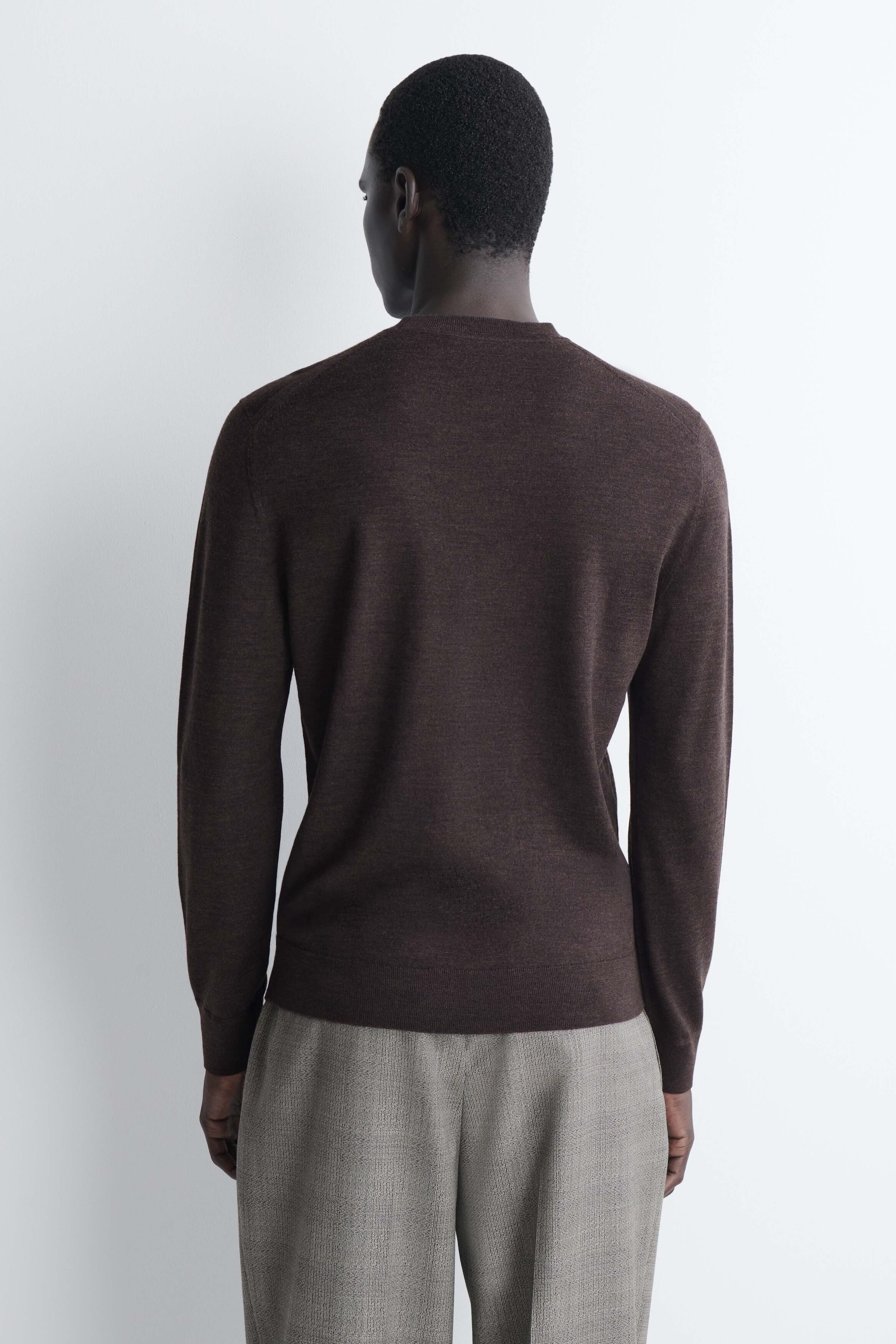 The slim merino wool crew-neck jumper in dark brown is shown from the back, highlighting its close-fitting silhouette and fine-knit texture. The long sleeves end in ribbed cuffs, and the ribbed hem rests neatly over light grey trousers. The slim merino wool crew-neck jumper in dark brown is shown from the back, highlighting its close-fitting silhouette and fine-knit texture. The long sleeves end in ribbed cuffs, and the ribbed hem rests neatly over light grey trousers.